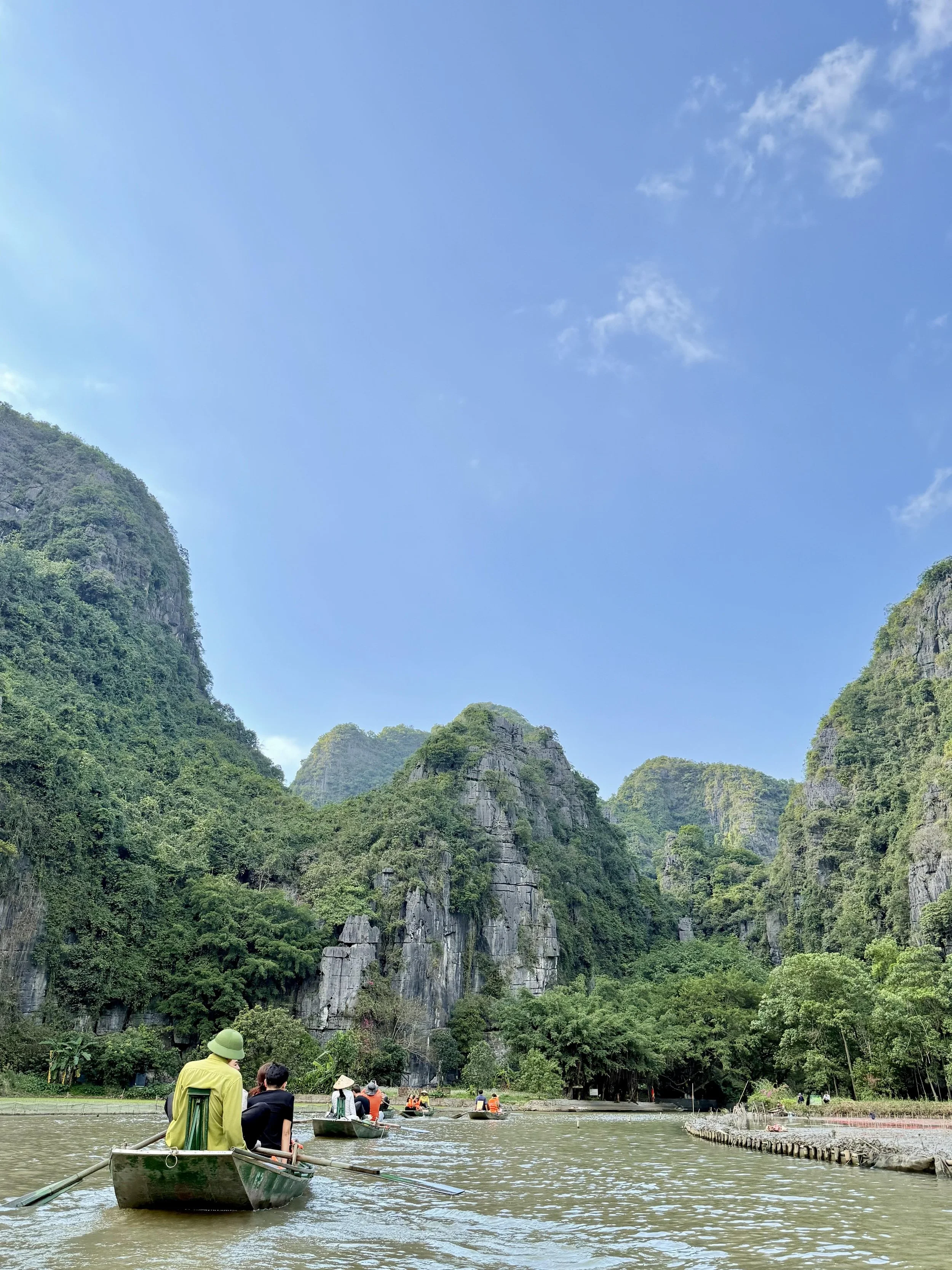 People in boats on a river surrounded by lush green mountains under a blue sky with a few clouds.