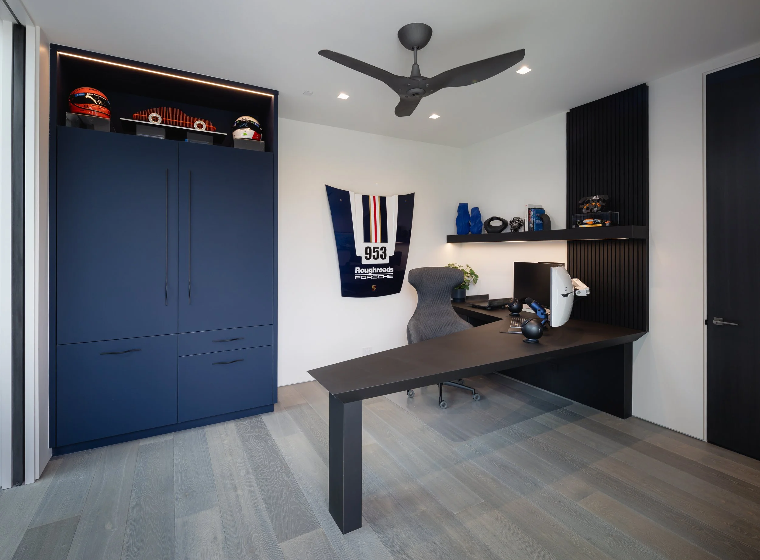 Modern home office with blue cabinet, racing flags, black desk, ergonomic chair, computer, and car-themed decor.