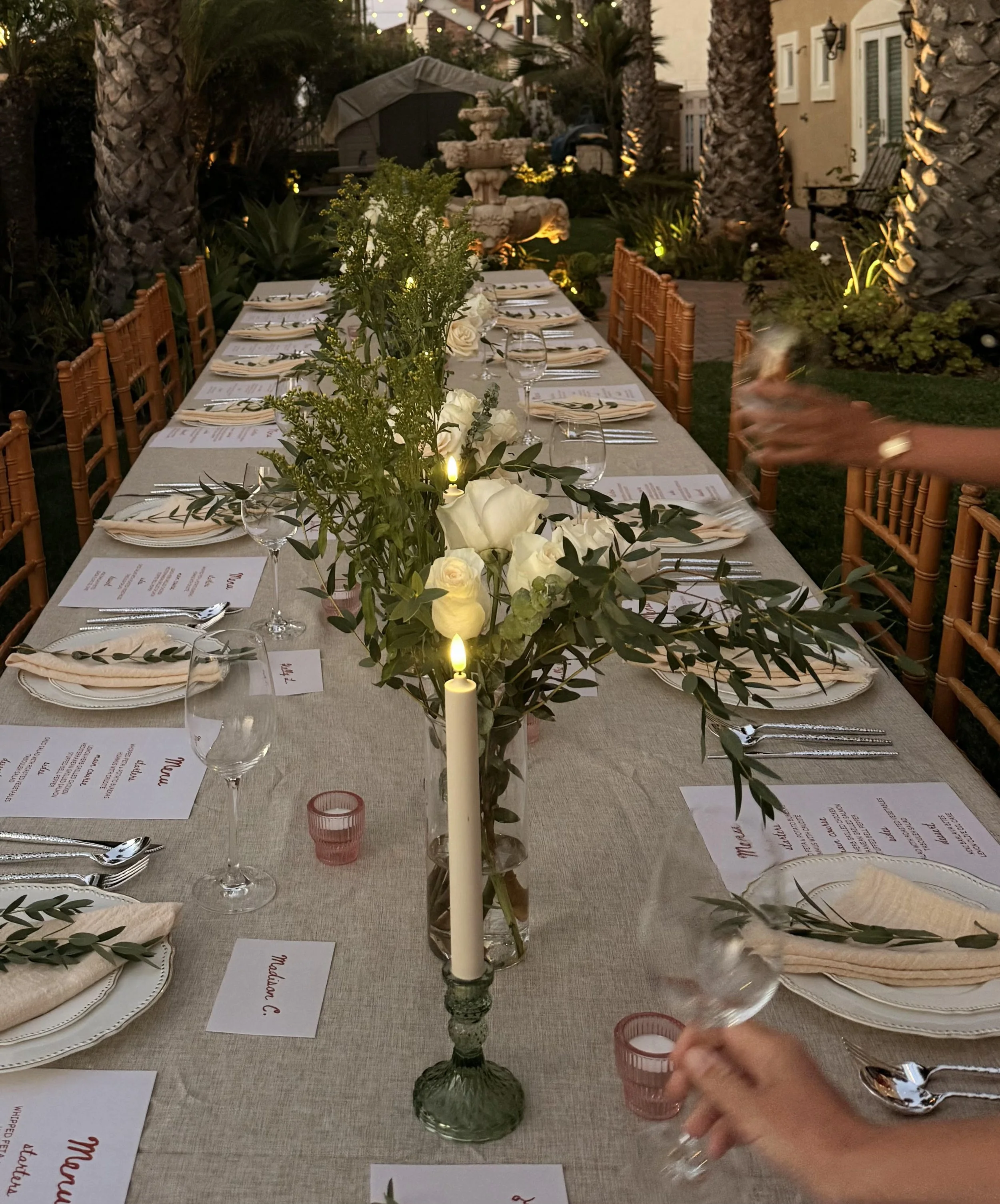 An outdoor dinner table decorated with white flowers, candles, and place settings, set for a formal event in the evening.