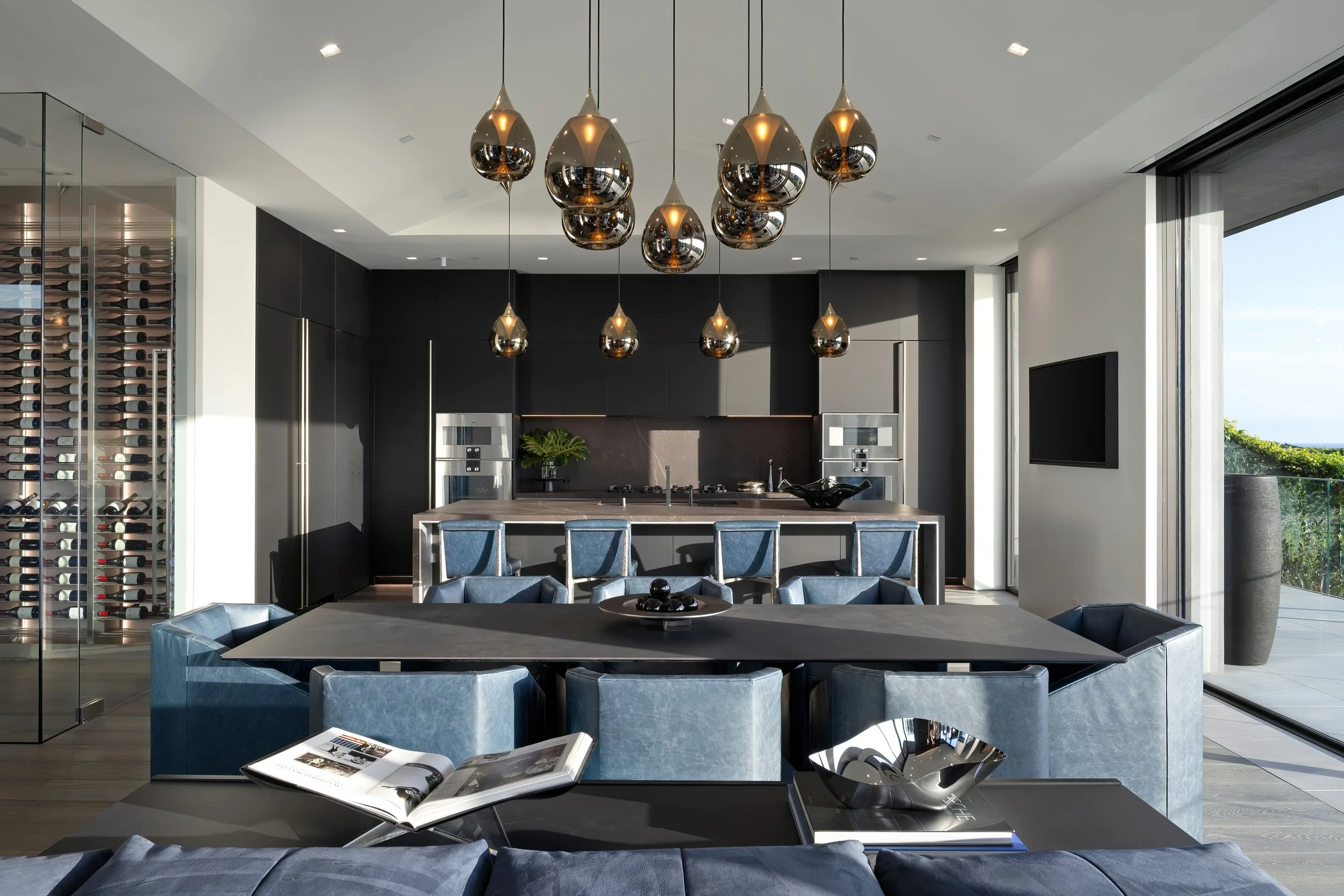 Modern kitchen and dining area with black cabinetry, stainless steel appliances, blue chairs, a black dining table, and decorative hanging pendant lights, with large windows showing an outdoor view.