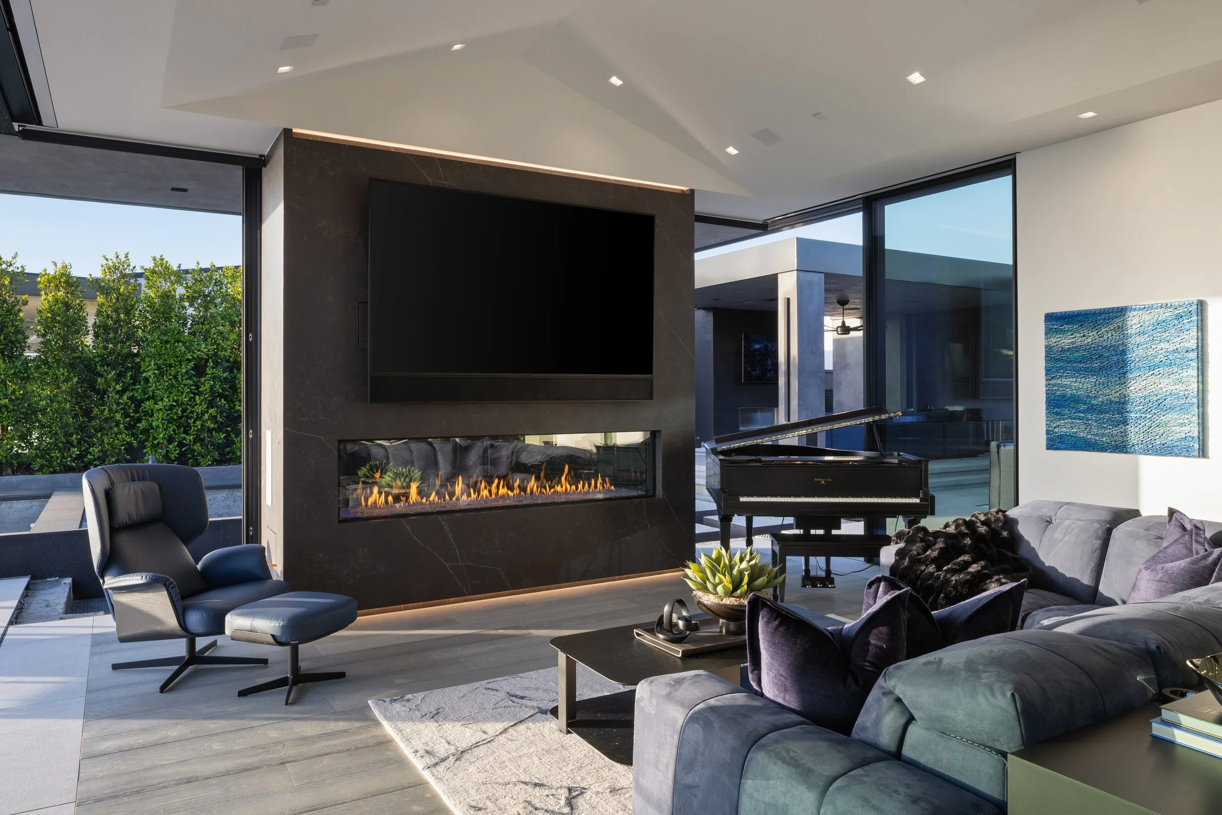 Modern living room with a black fireplace, flat-screen TV, grand piano, gray sofa, black armchair with matching footstool, blue abstract wall art, and large windows showing greenery outside.