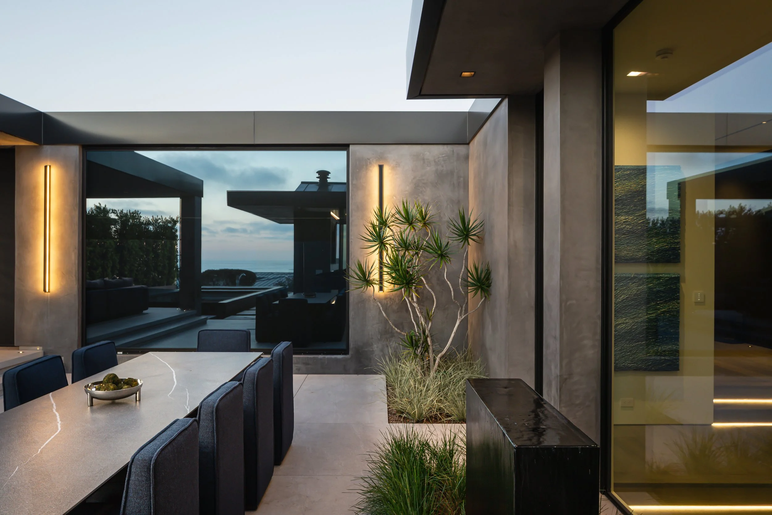 Modern outdoor patio with a dining table, black chairs, a large potted plant, and a view of the ocean through glass walls.