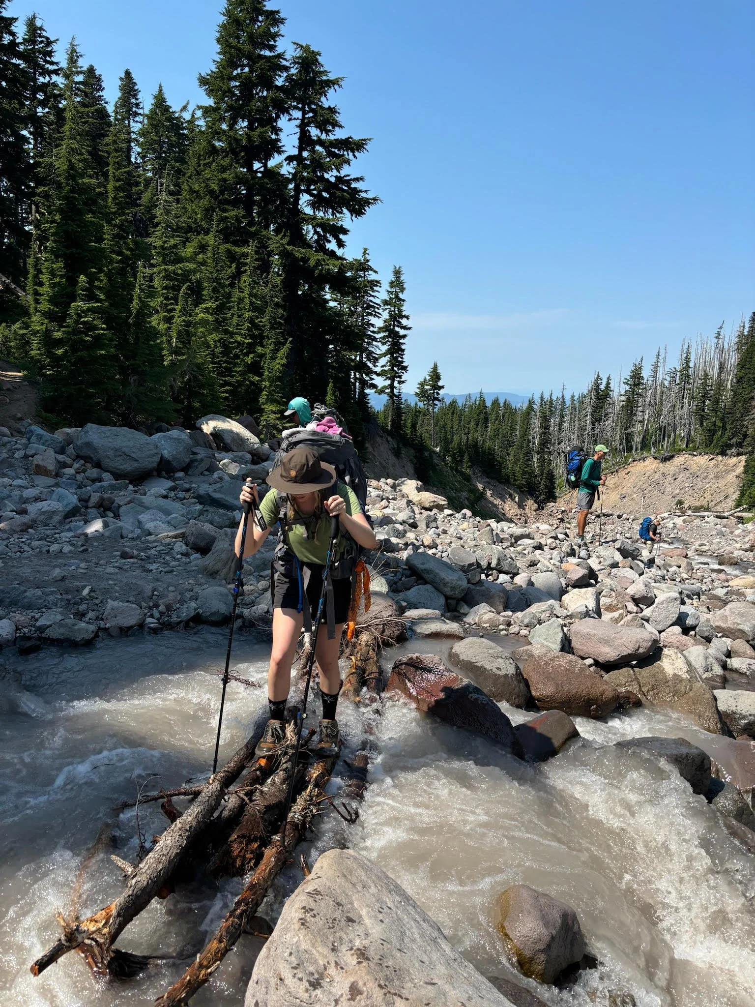 A group of hikers crossing a rushing mountain stream on fallen logs, surrounded by trees and rocky terrain under a clear blue sky.
