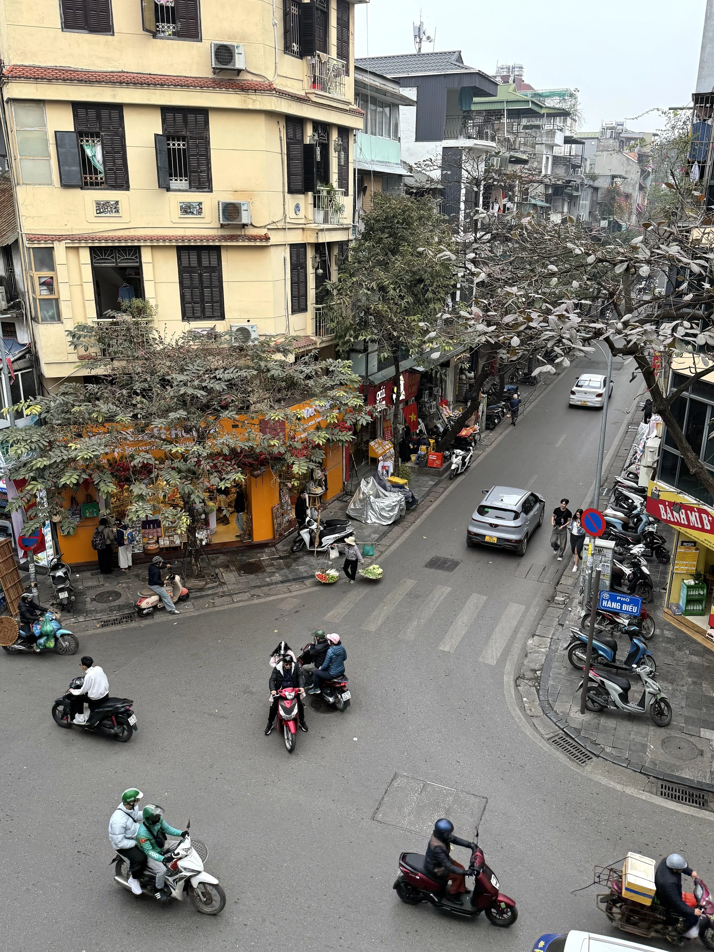 City street scene with motorcycles, cars, pedestrians, shops, and buildings in an urban area with trees.