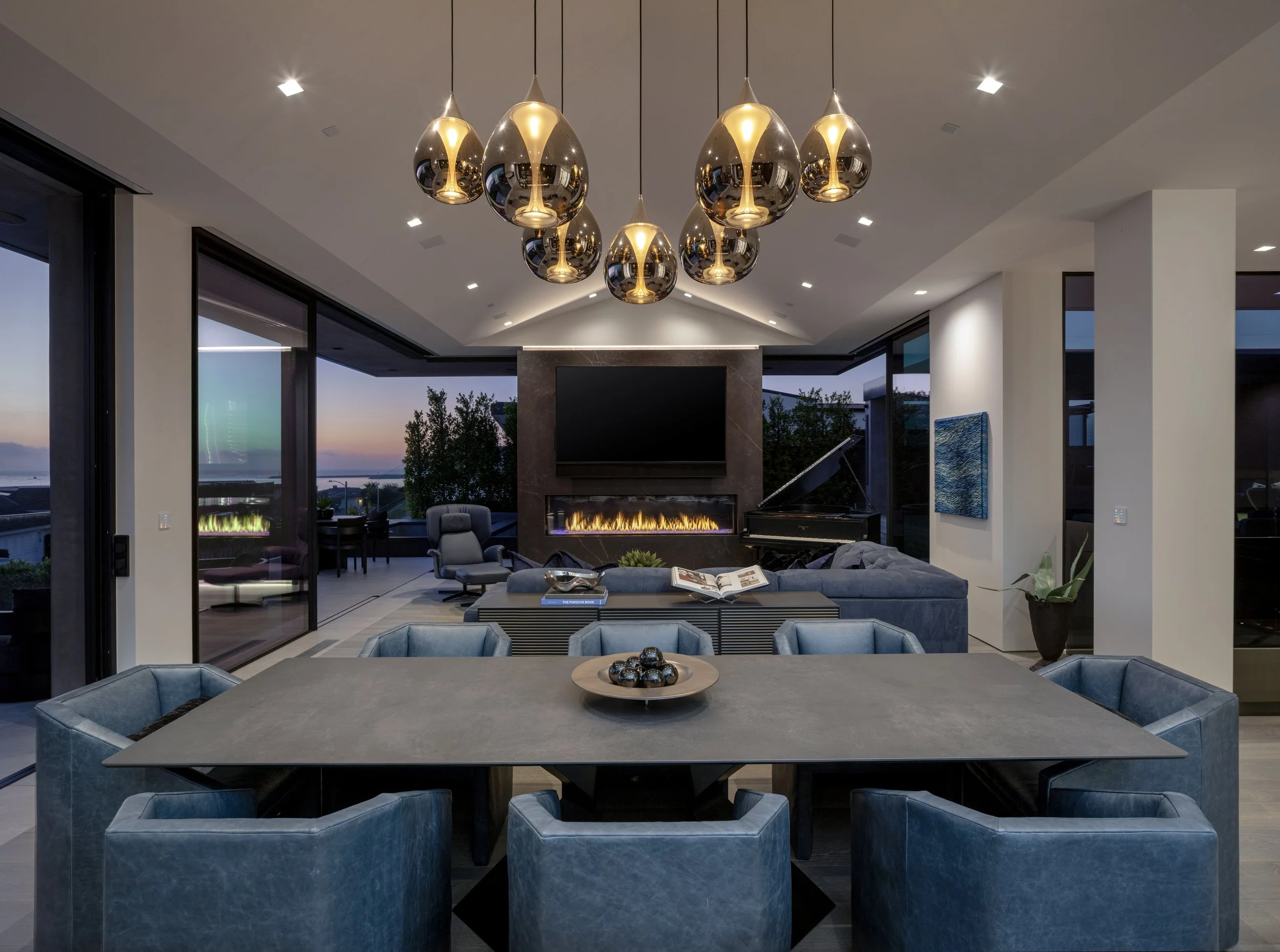 Modern dining room with a large gray table surrounded by blue upholstered chairs, a living area with a fireplace, a flat-screen TV on the wall, a grand piano, and large windows showing outdoor seating and sunset view.
