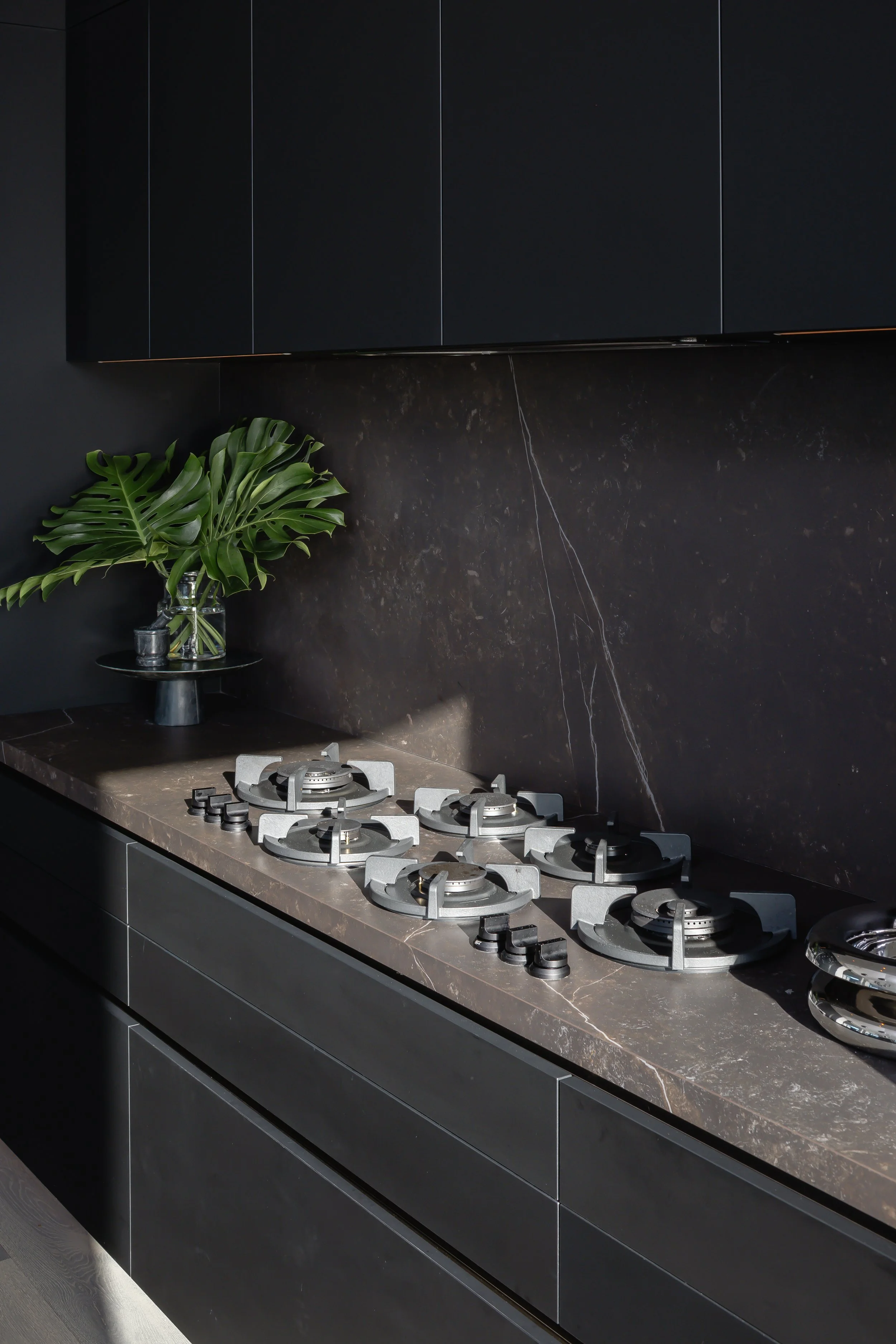 Modern black kitchen with a marble countertop, gas stove with six burners, a black and dark grey cabinet, and a glass vase with large green monstera leaves.