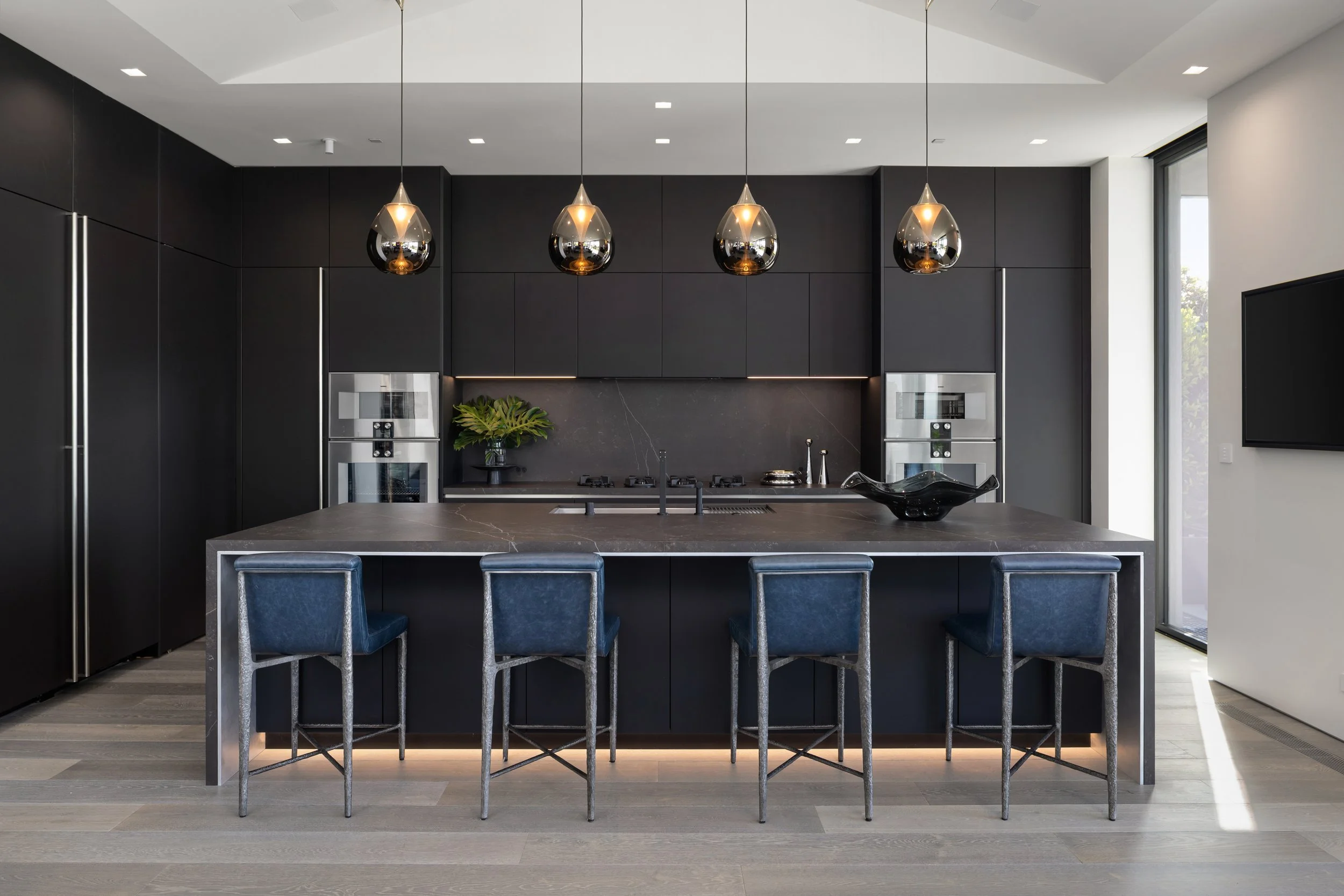 Modern black kitchen with central island, four blue bar stools, black pendant lights, built-in stainless steel appliances, potted plant, black bowl, and a large window with a sliding door.
