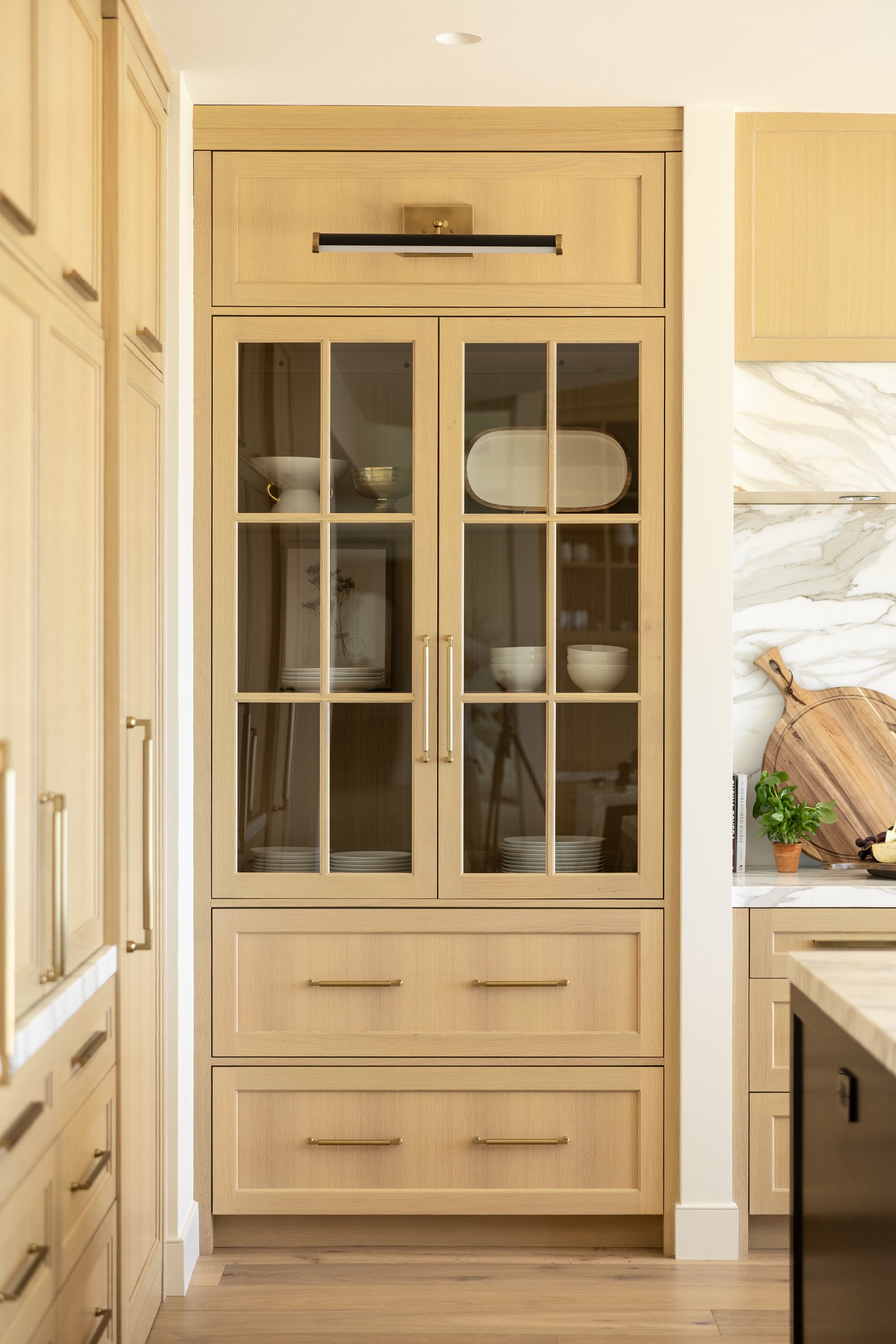 A light wood kitchen cabinet with glass-paneled doors displaying white and beige dishes, and three drawers below with gold handles, situated against a white wall with a marble backsplash and a cutting board with herbs on the counter.