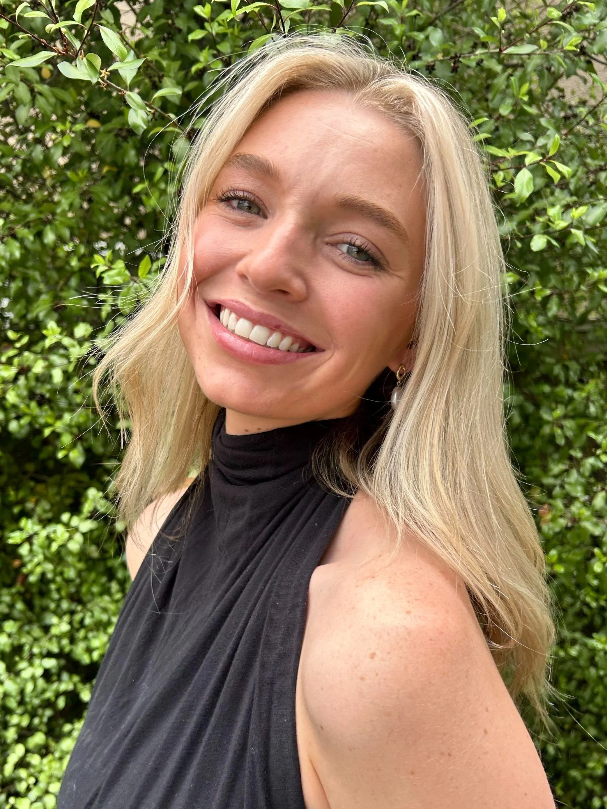 Close-up of a smiling young woman with blonde hair, wearing a black sleeveless top, standing outdoors in front of a green leafy background.