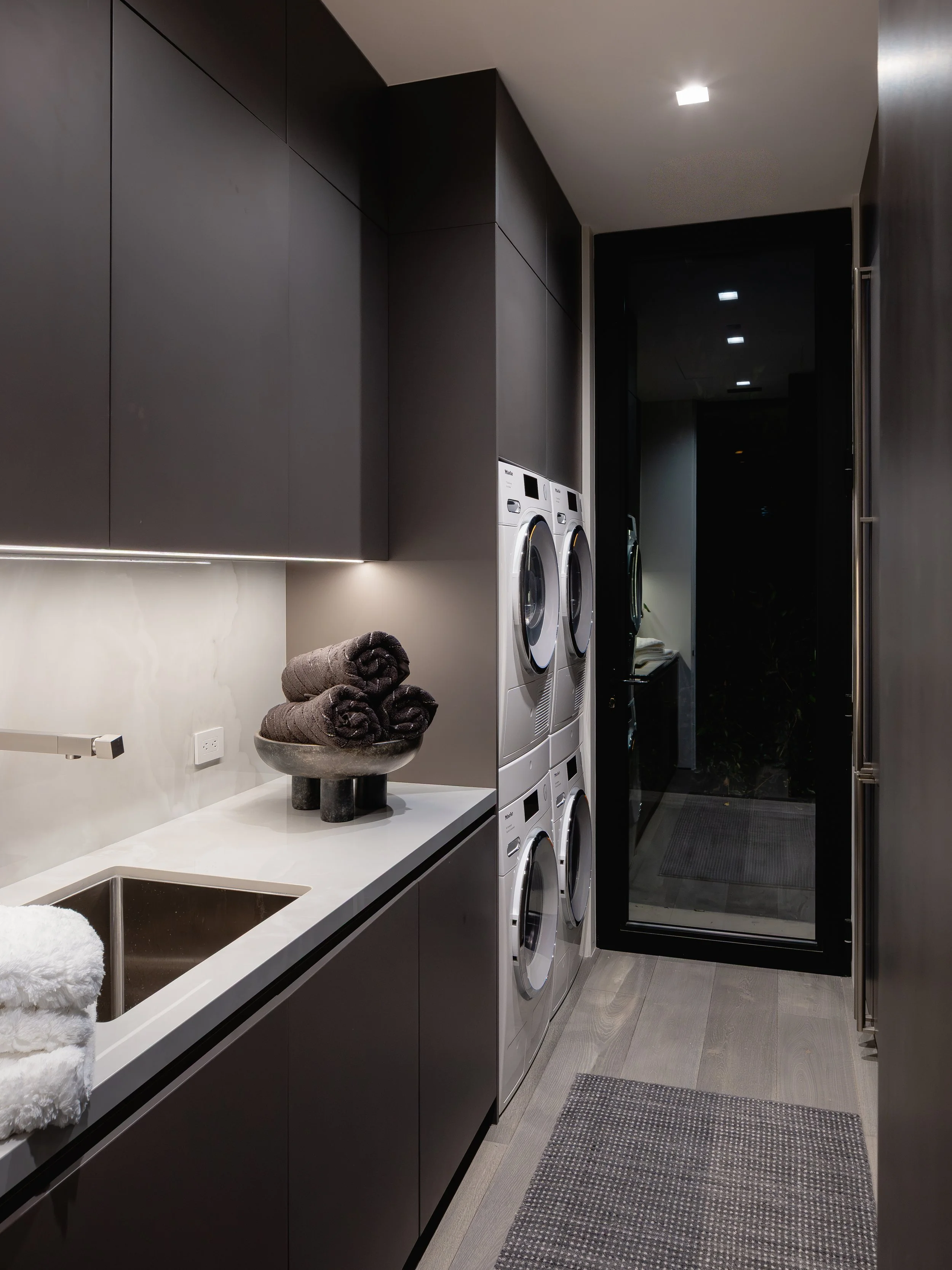 Modern laundry room with black cabinets, stacked washing machine and dryer, white countertops, rolled towels, and a door leading outside.