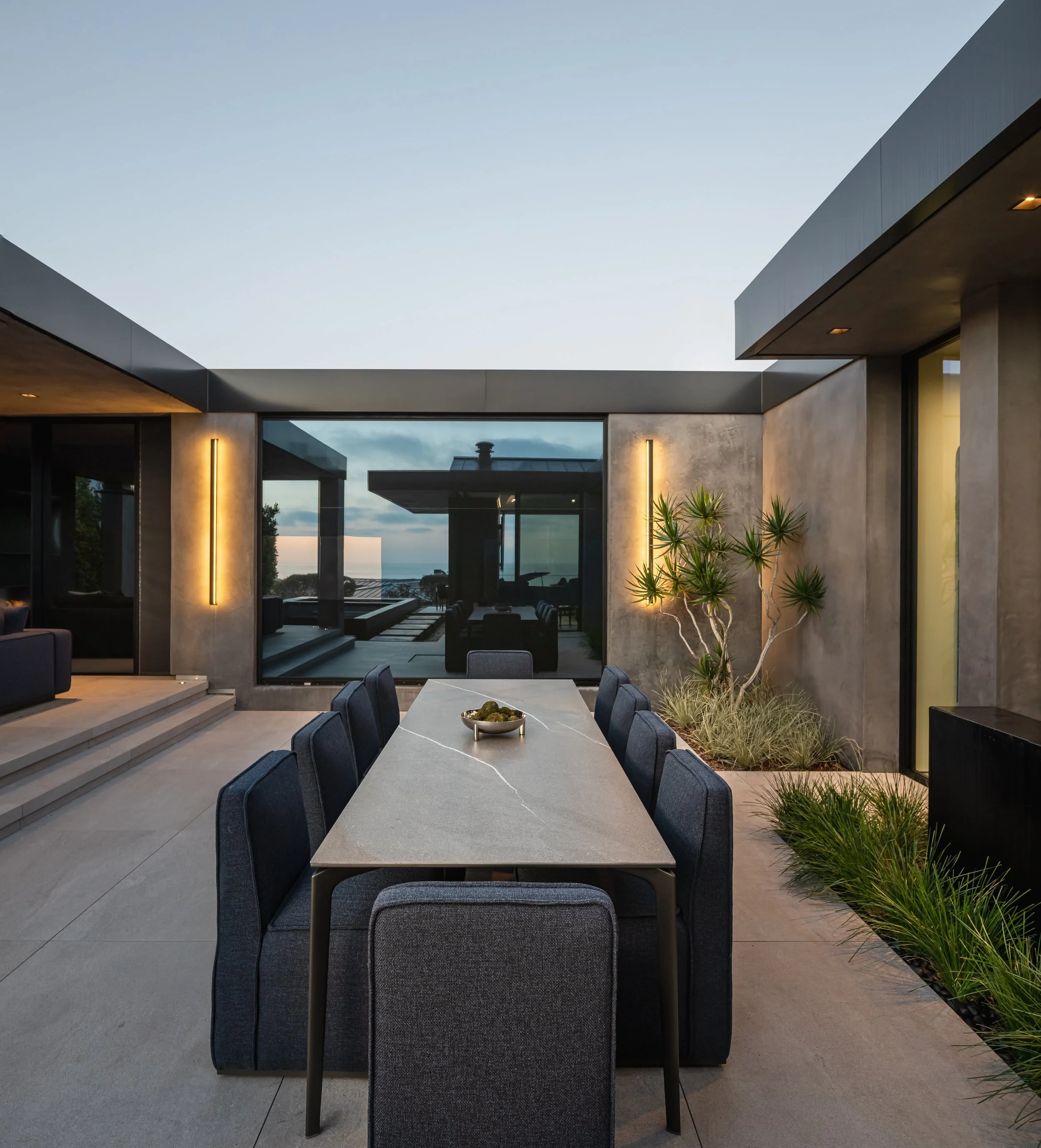 Modern outdoor patio with a long dining table, 8 chairs, plants, and large glass doors leading to an indoor space, during dusk.