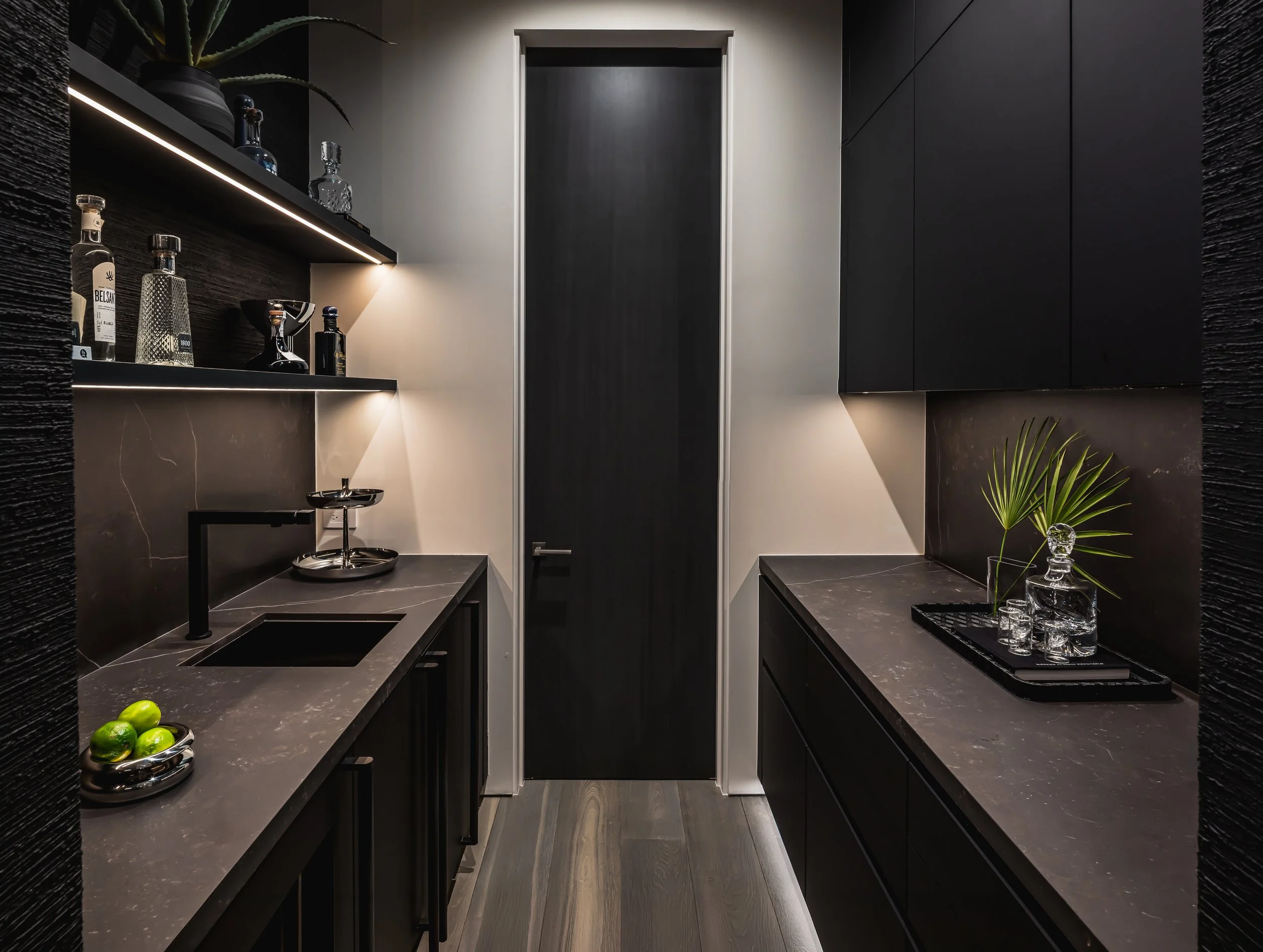 A modern, black and gray kitchen with black cabinets, gray countertops, and decorative items including bottles, glassware, and a small plant.