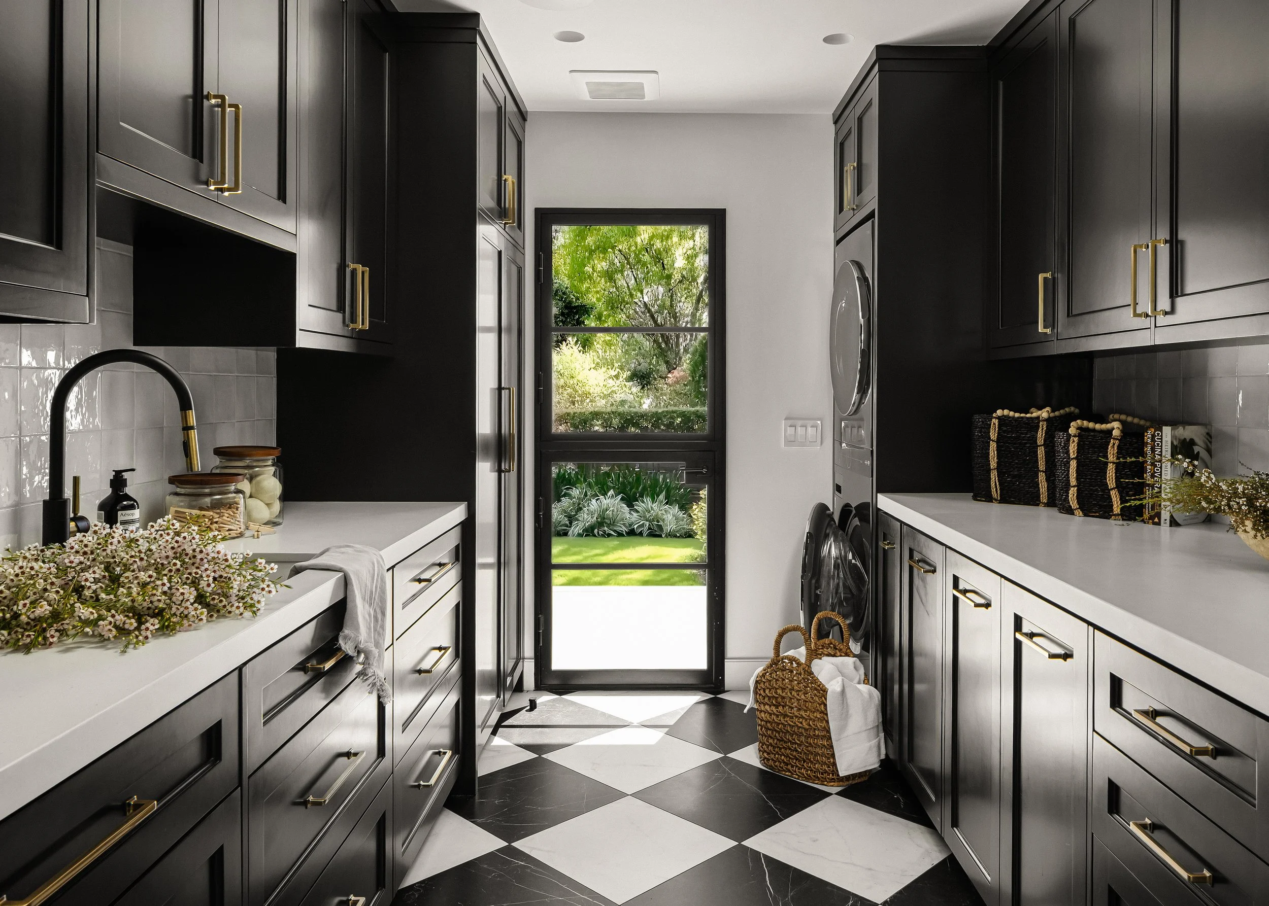 Modern black kitchen with white countertops, gold hardware, a window showing trees outside, and black-and-white checkered floor.