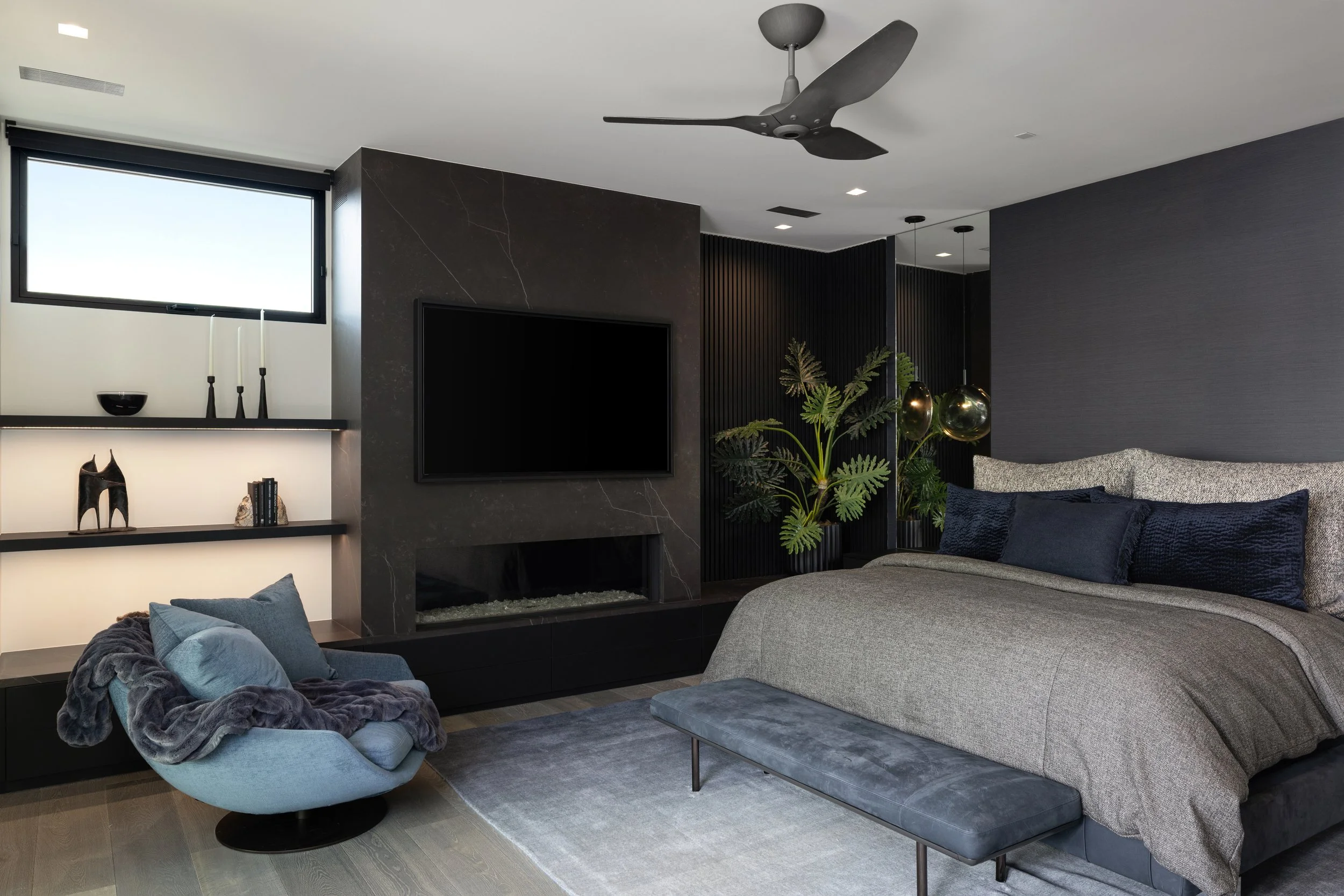 Modern bedroom with a large bed, gray bedding, black pillows, and a gray bench at the end. A flat-screen TV is mounted on a dark marble fireplace wall. There is a black ceiling fan, a window, a built-in shelf with decor items, and a black accent wall