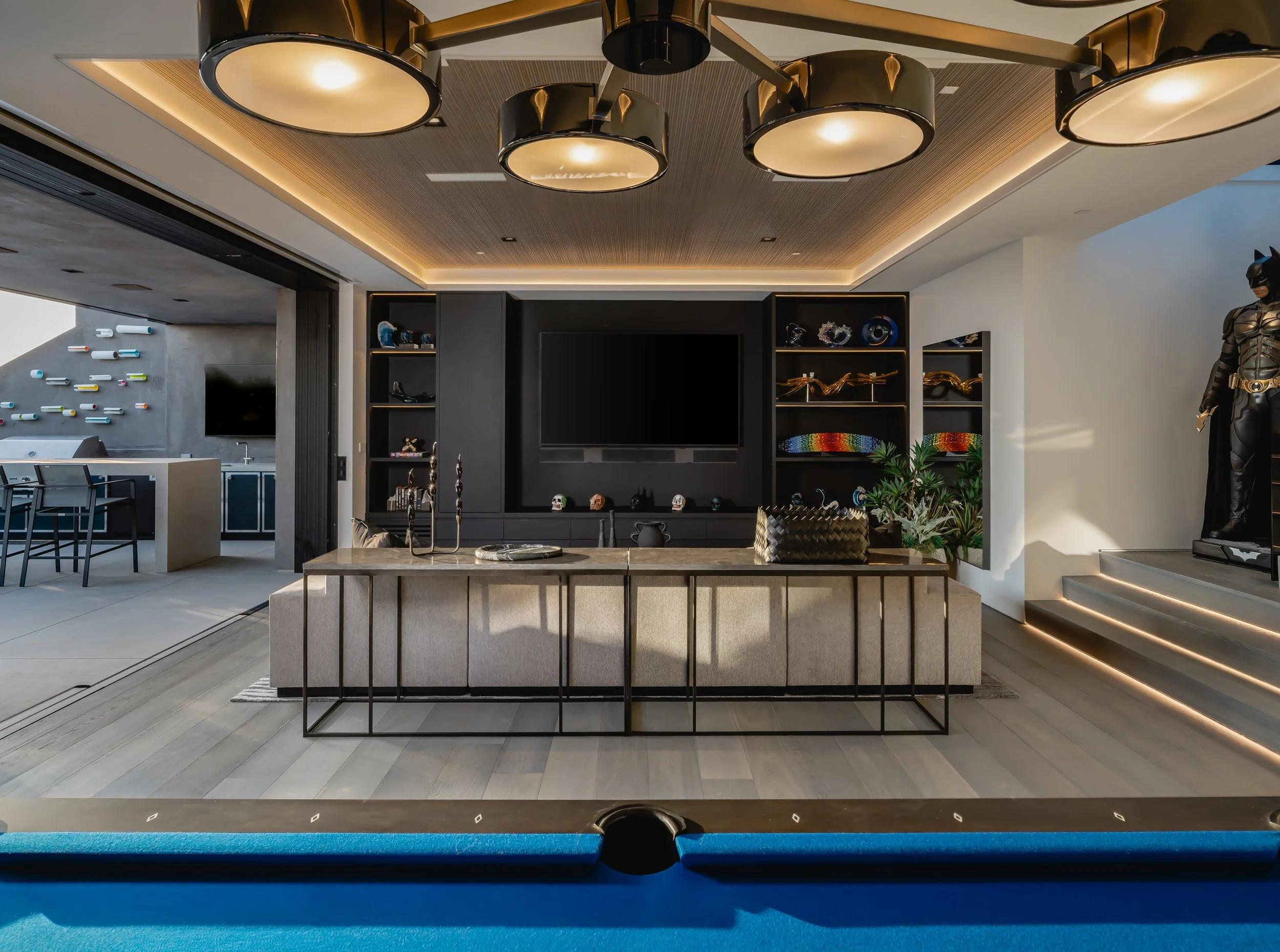 Modern living room with a large black entertainment center, shelves with decorative objects, a beige sofa, a small table, and a staircase with LED lighting. Part of a pool table is visible at the bottom of the image.