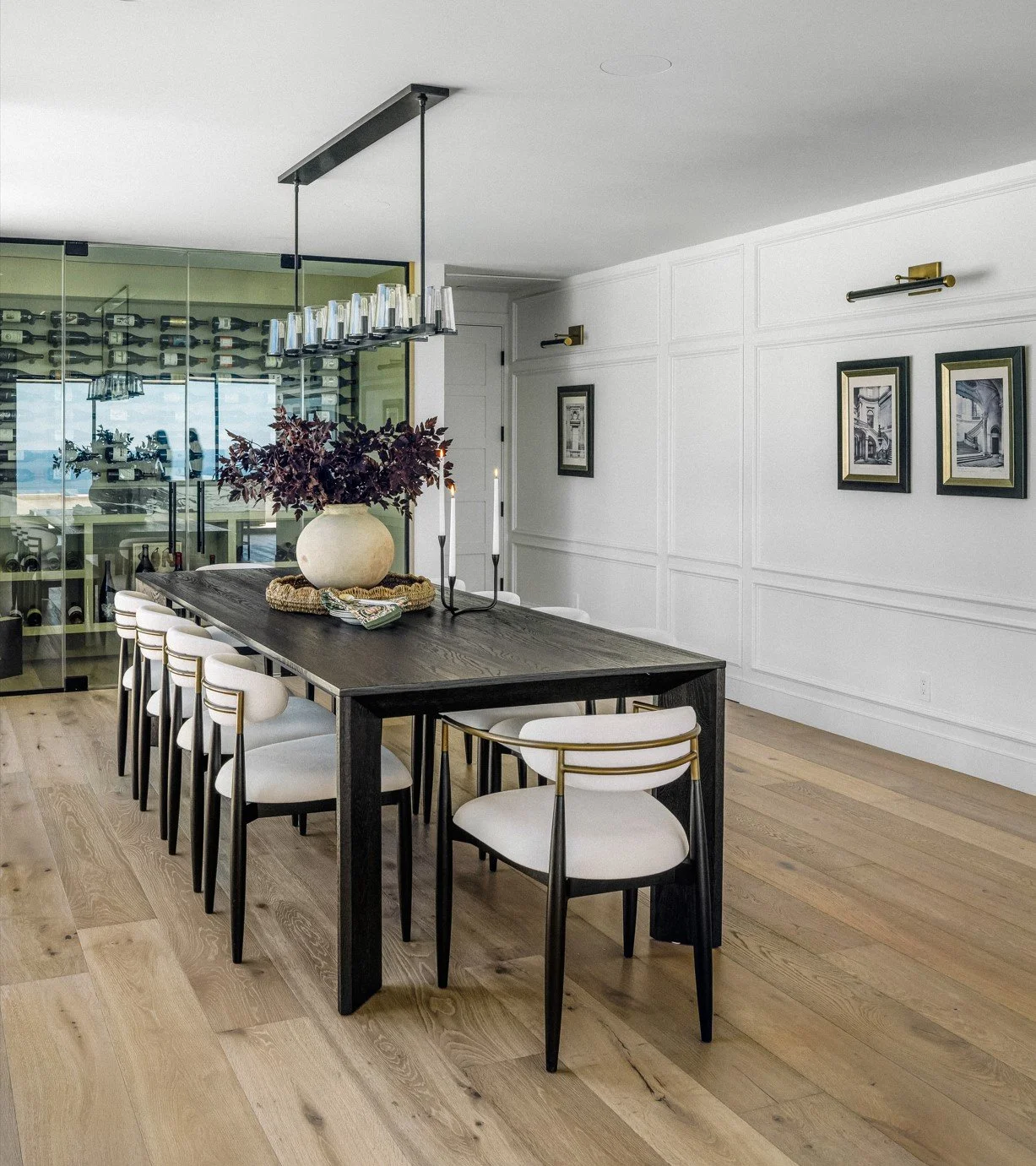 Modern dining room with a large wooden table, white and gold chairs, a large vase with purple foliage, and a wine cellar visible through glass walls.