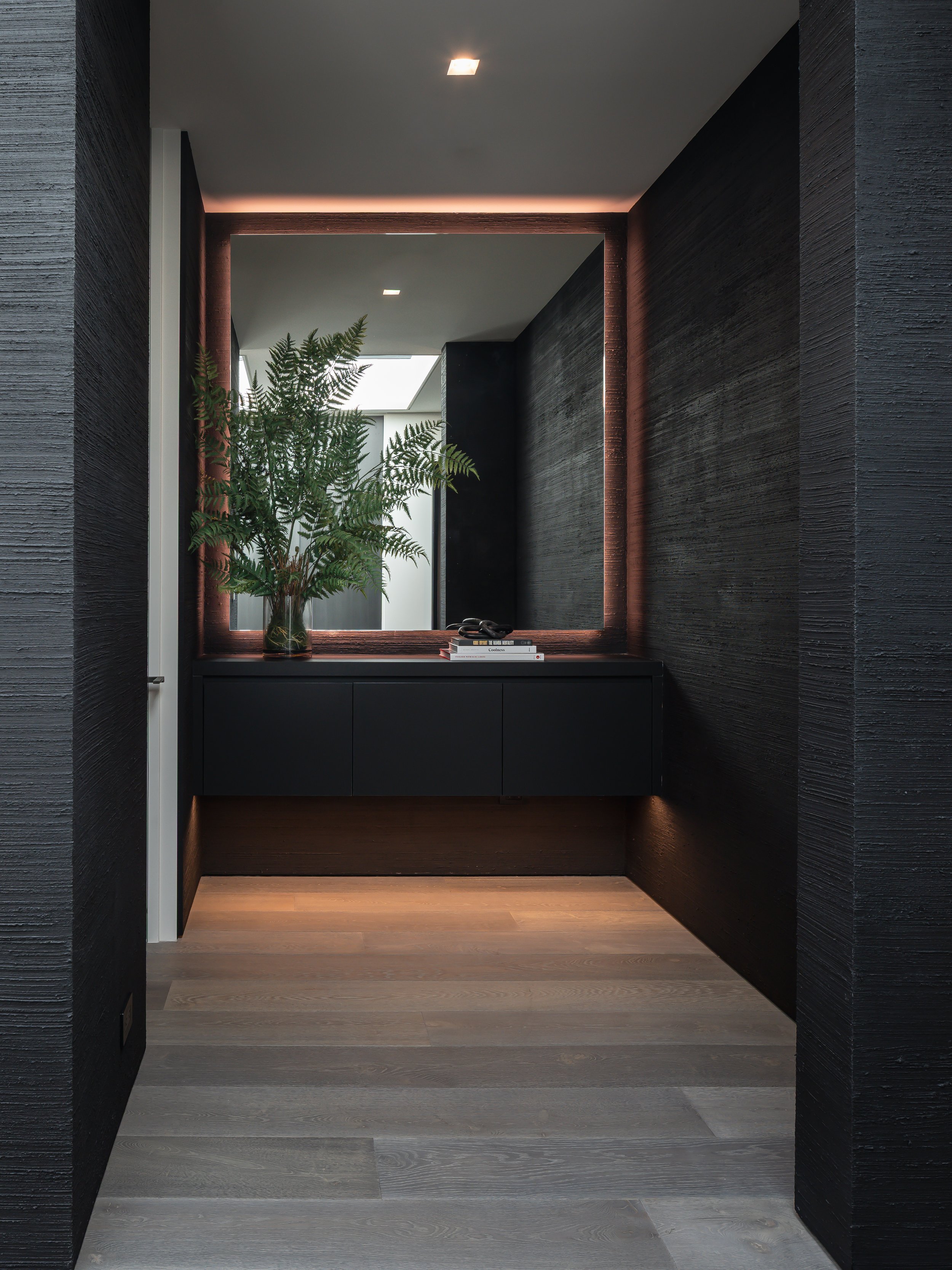 A modern entryway with black textured walls, a large mirror with a copper frame, a black dresser, a potted fern, and a stack of books and sunglasses on top, illuminated with concealed lighting.