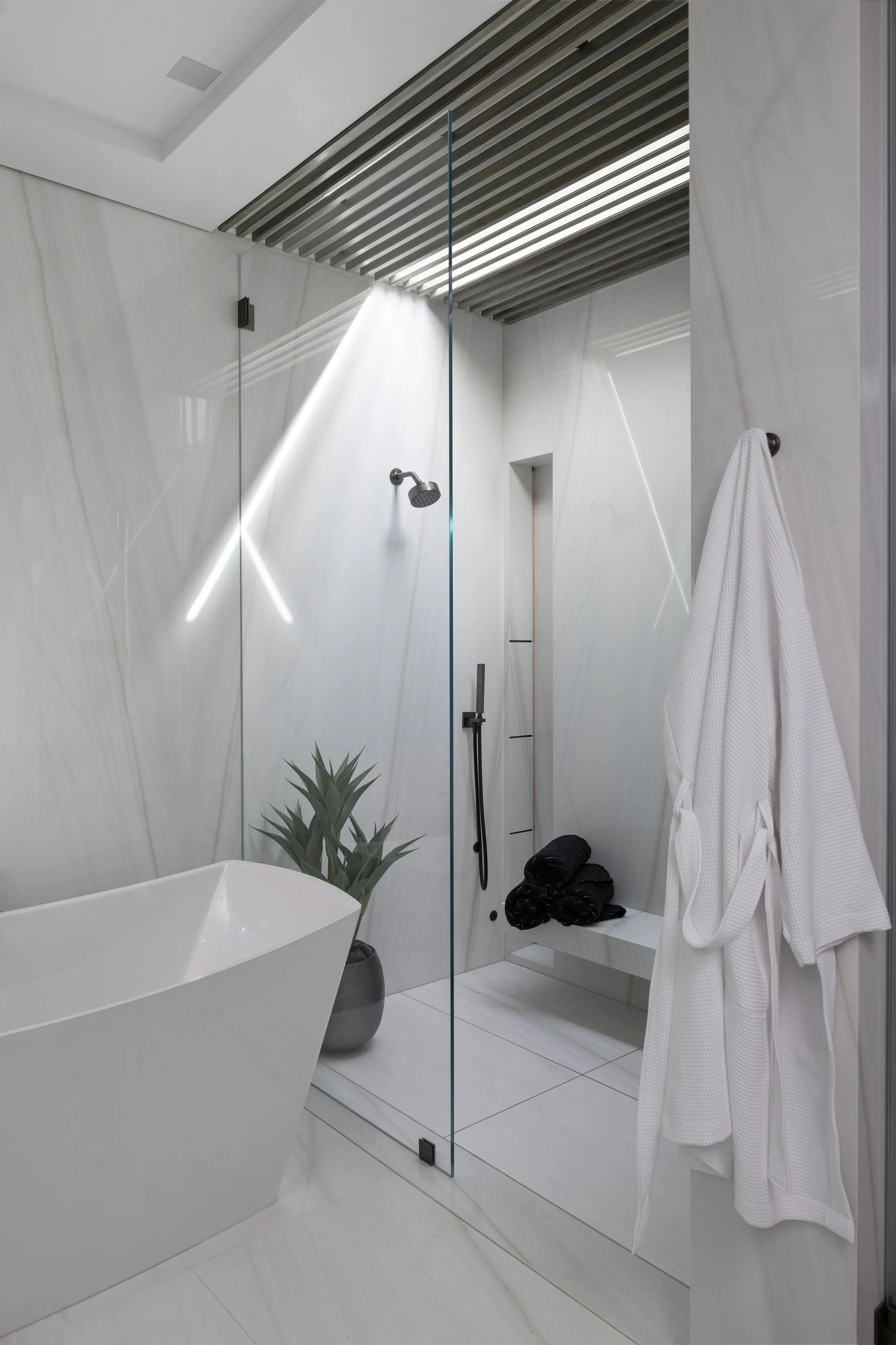 Modern bathroom with a free-standing bathtub, a glass-enclosed shower area, white marble walls and floor, a potted plant, and a white bathrobe hanging on the wall.