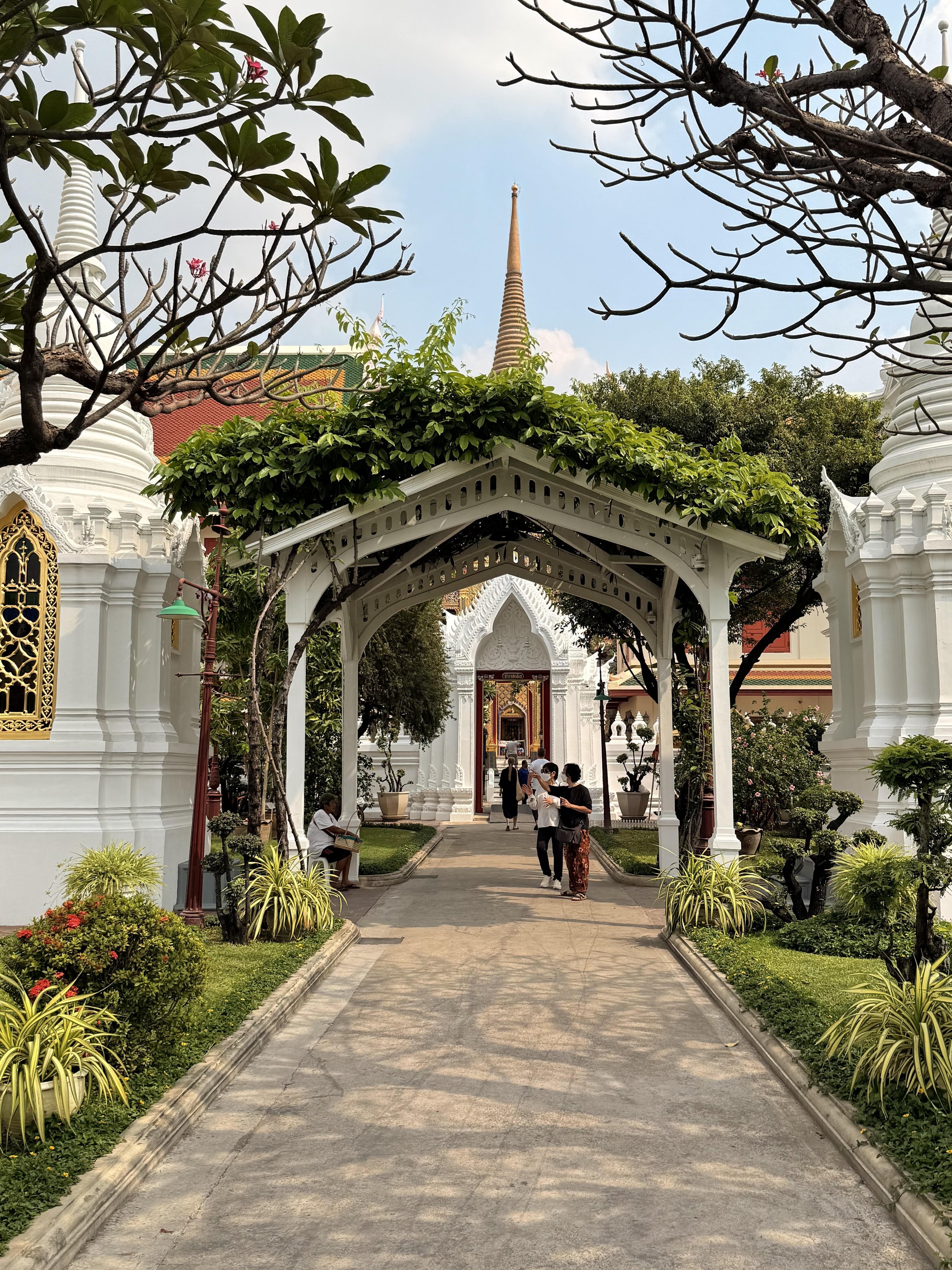 View of a pathway leading to a temple, with people walking and sitting, surrounded by greenery and ornate white structures.