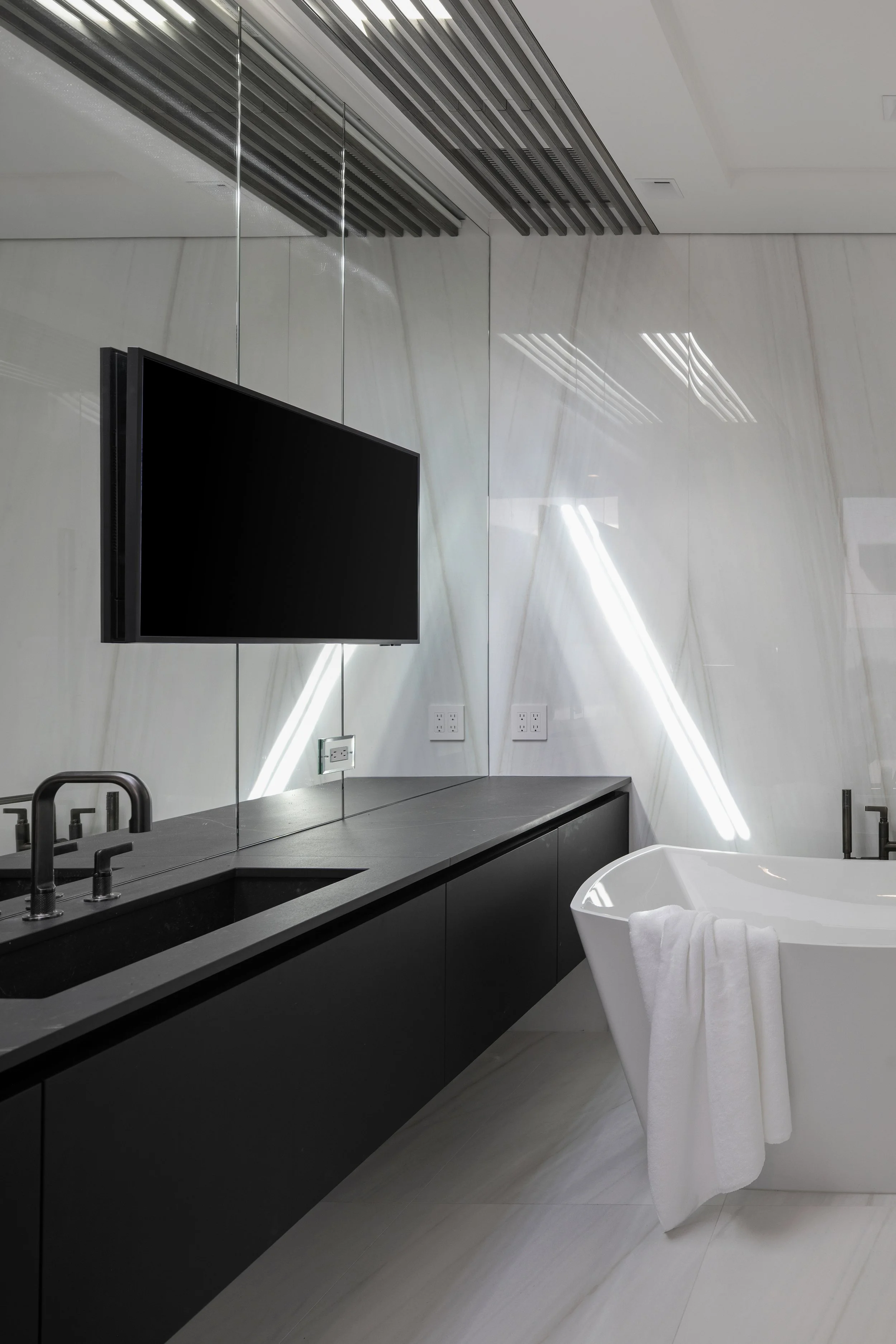 Modern bathroom with a white bathtub, a black countertop with a sink, a flat-screen TV mounted on a mirror, and a large mirror reflecting the ceiling lights.