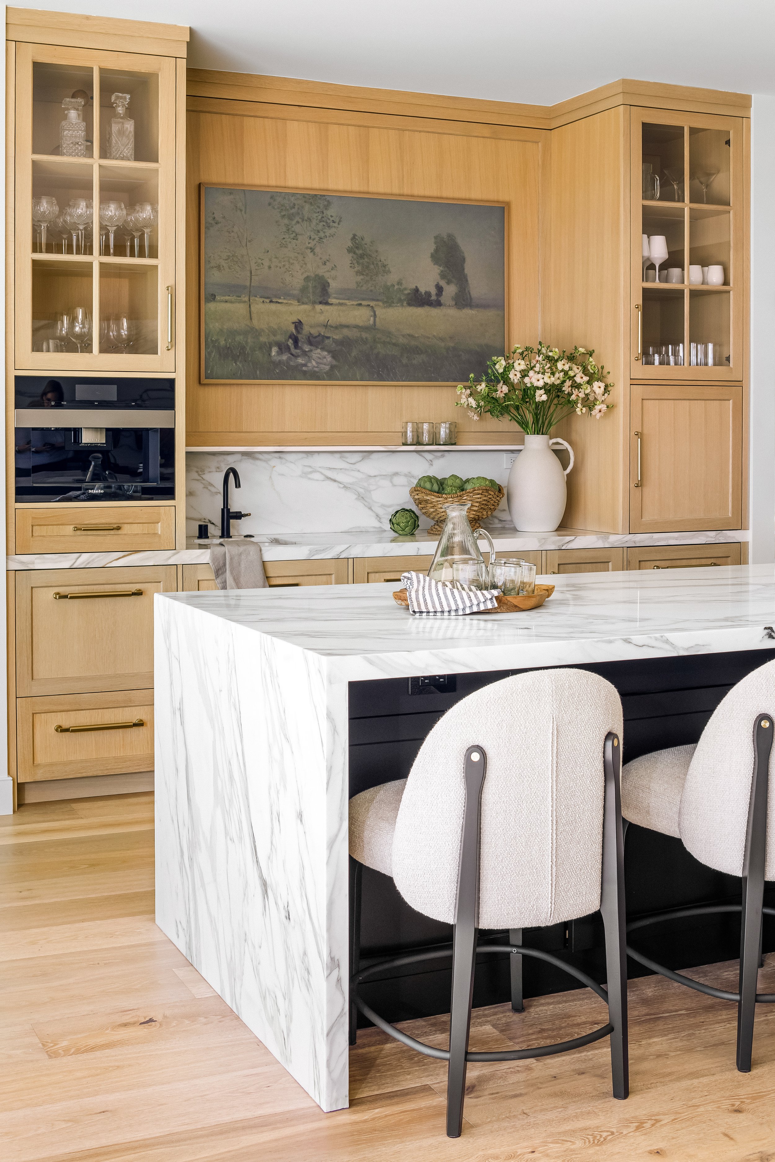 A modern kitchen featuring light wood cabinets with gold handles, a white marble island with gray veining, and a backsplash. There are two glass-front cabinets displaying glassware, a built-in microwave, and a sink with a black faucet. Decor includes