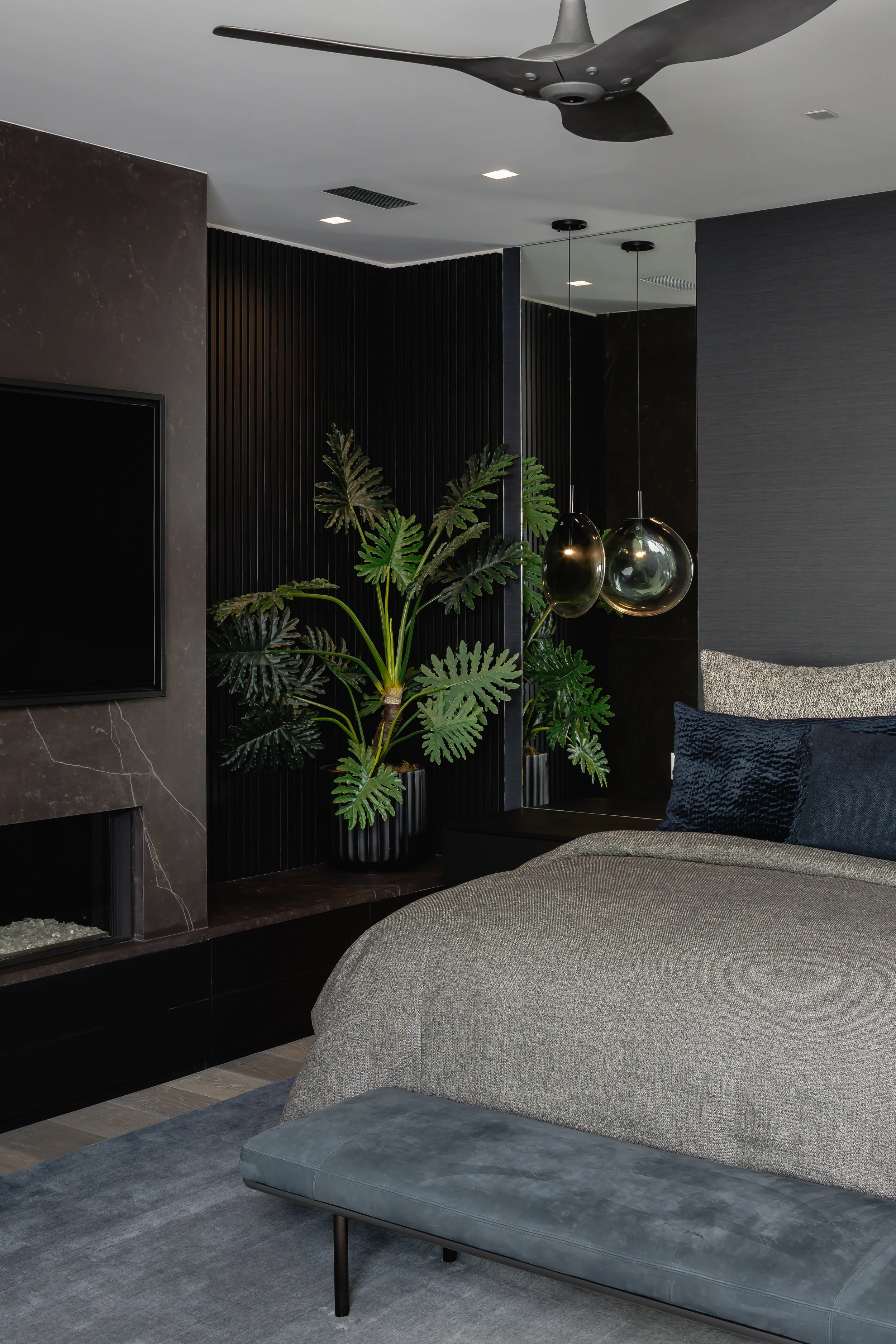 Modern bedroom with dark walls, a large green plant in a black pot, a flat screen TV, pendant lights, gray and black bedding, and a ceiling fan.