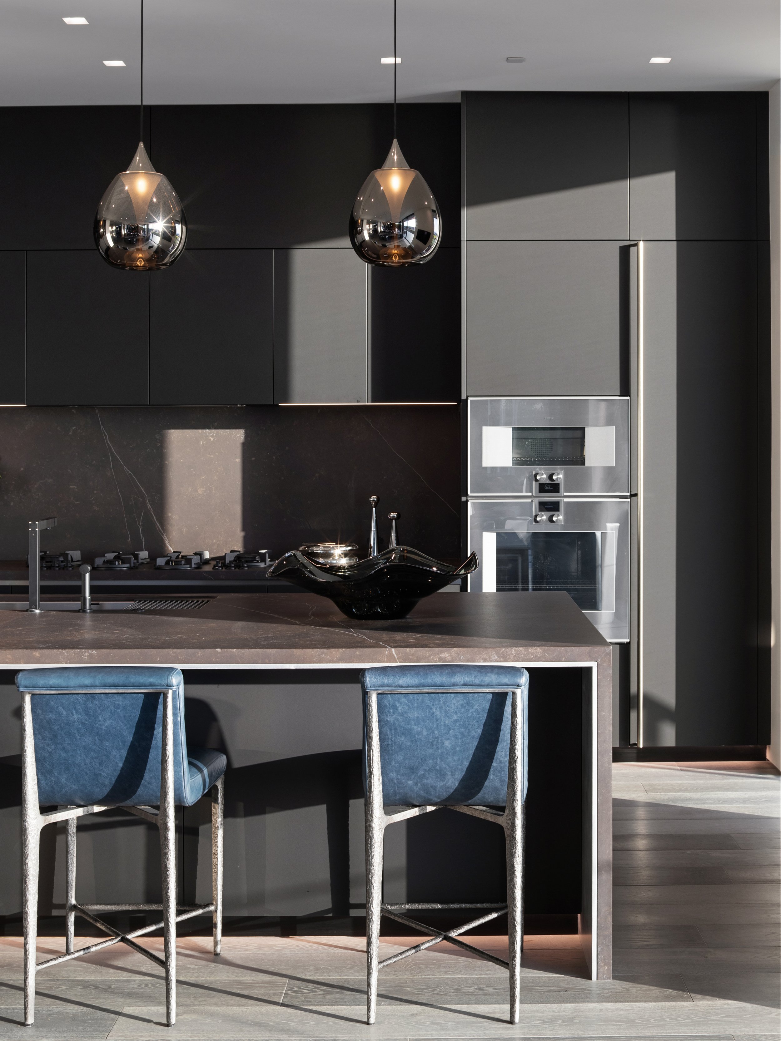 Modern kitchen with black cabinets, a marble island with two blue bar stools, and a stainless steel oven and microwave. Two black pendant lights hang above the island.