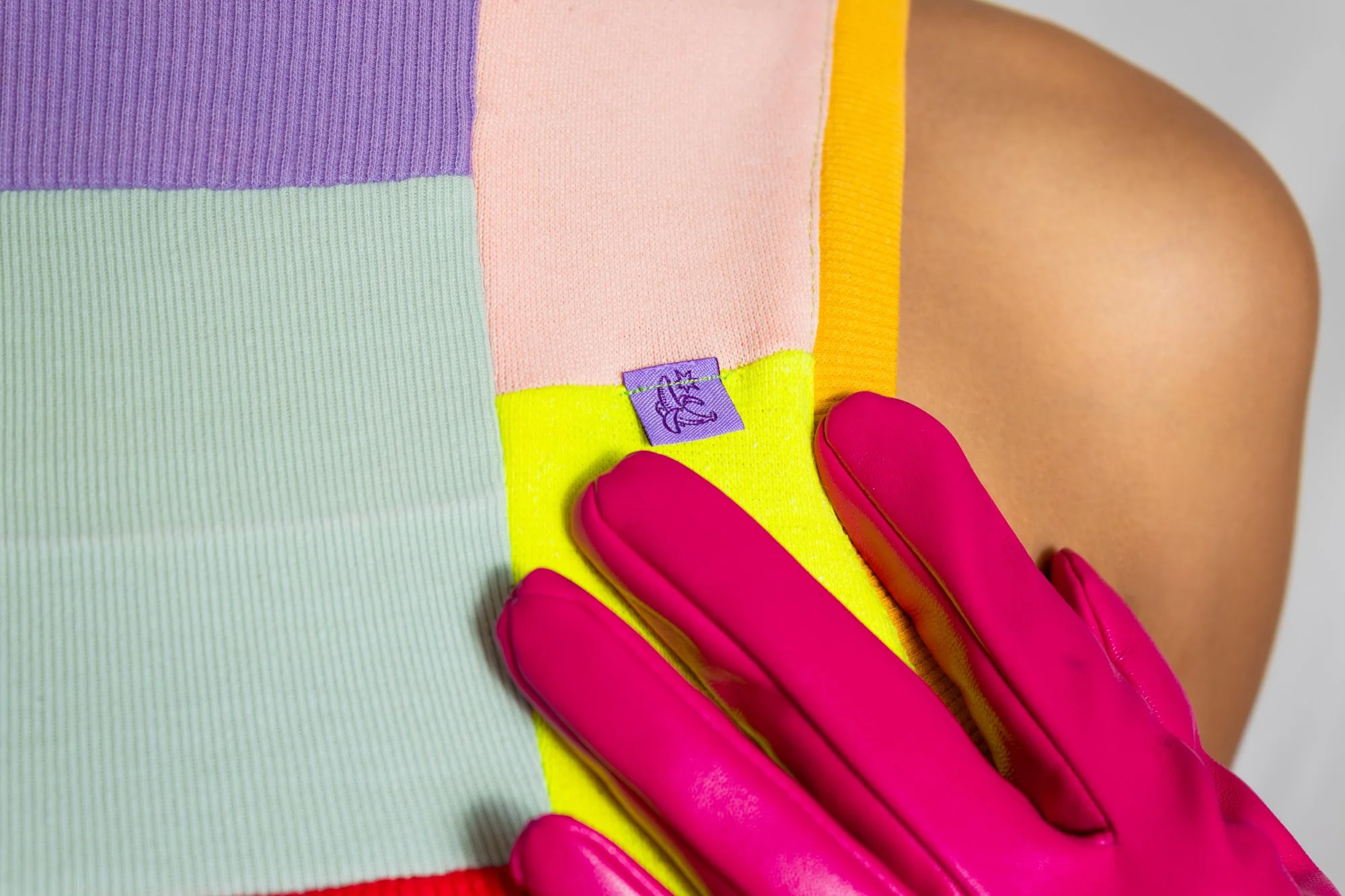 Close-up of a person wearing a colorful, striped shirt and a pink glove, touching their shoulder with their hand, showing a small purple tag on the shirt.