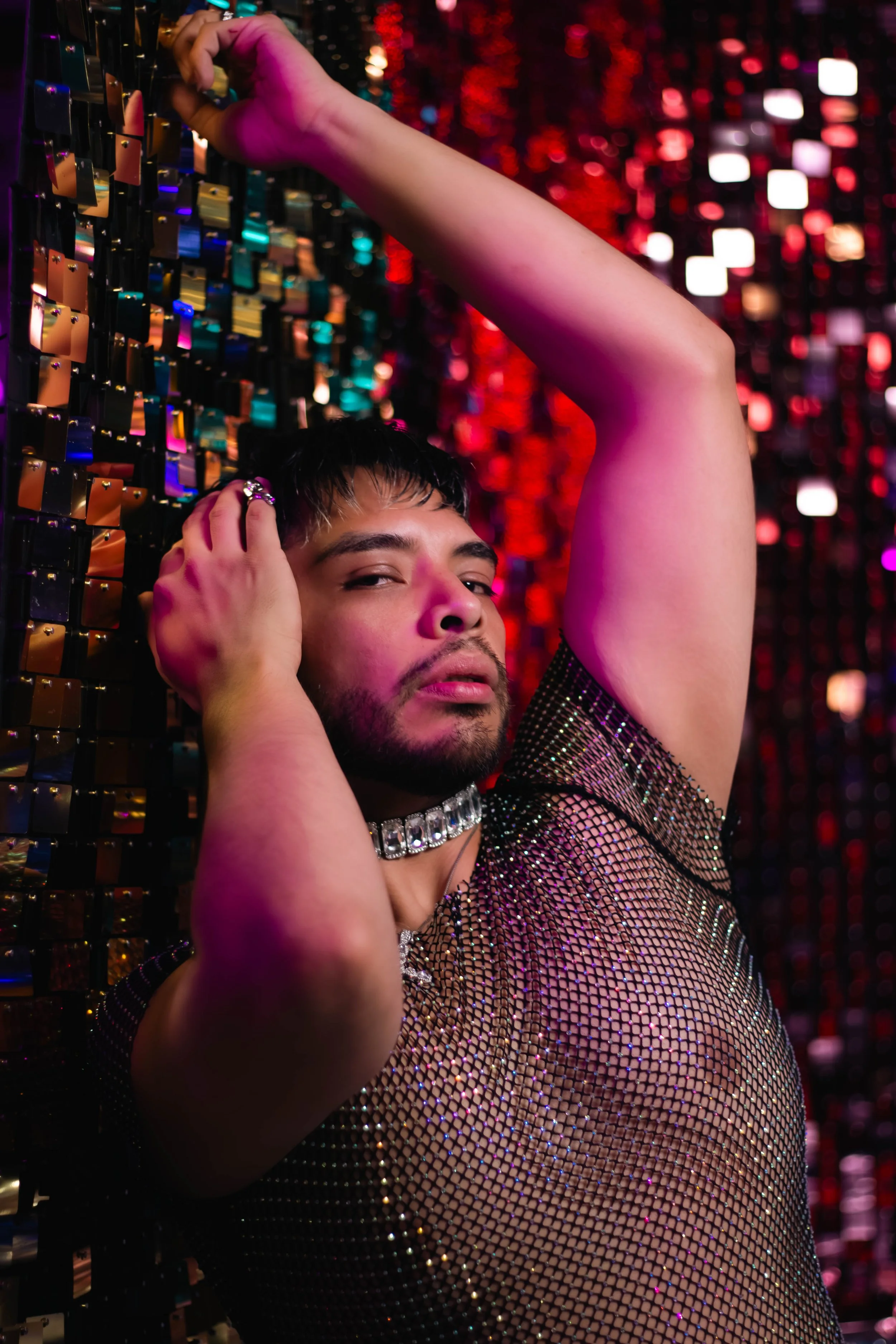 Person with short dark hair and beard, wearing a sparkly mesh top and rhinestone choker, With a colorful, reflective background, striking a pose with one arm raised and hand on head.