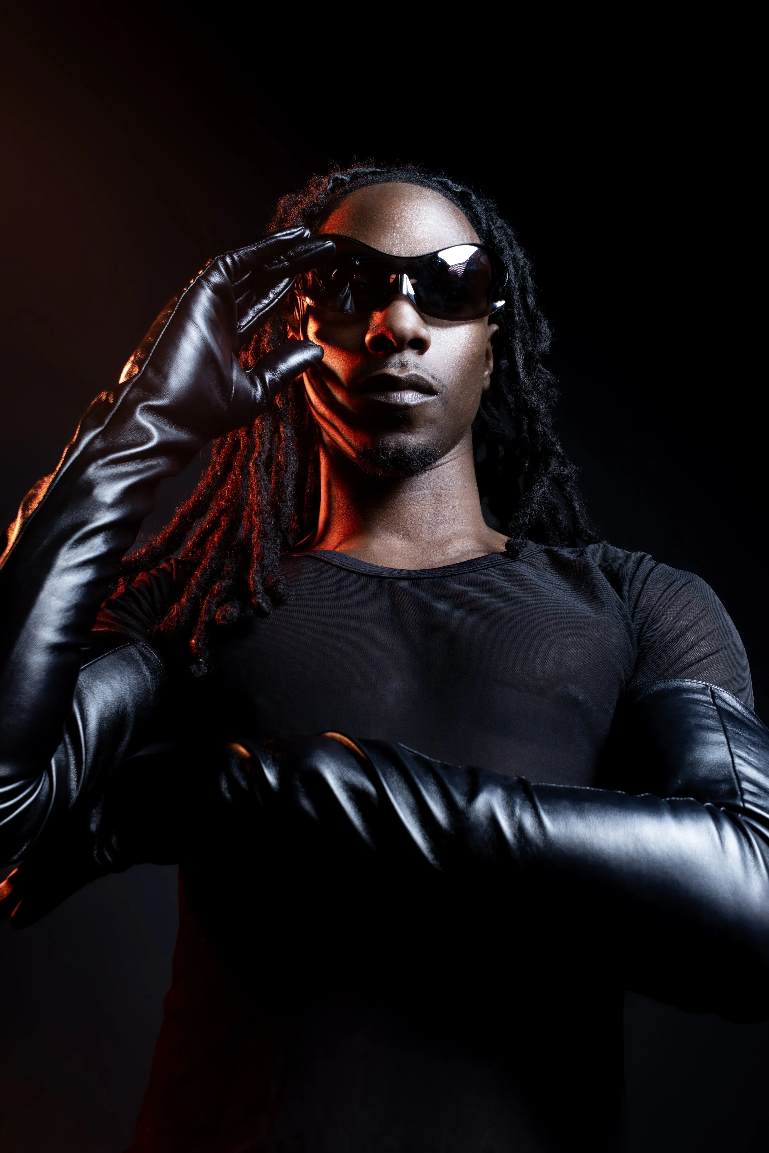 A person with dreadlocks wearing black sunglasses, a black top, and black leather gloves, standing against a dark background with red and orange lighting highlights.
