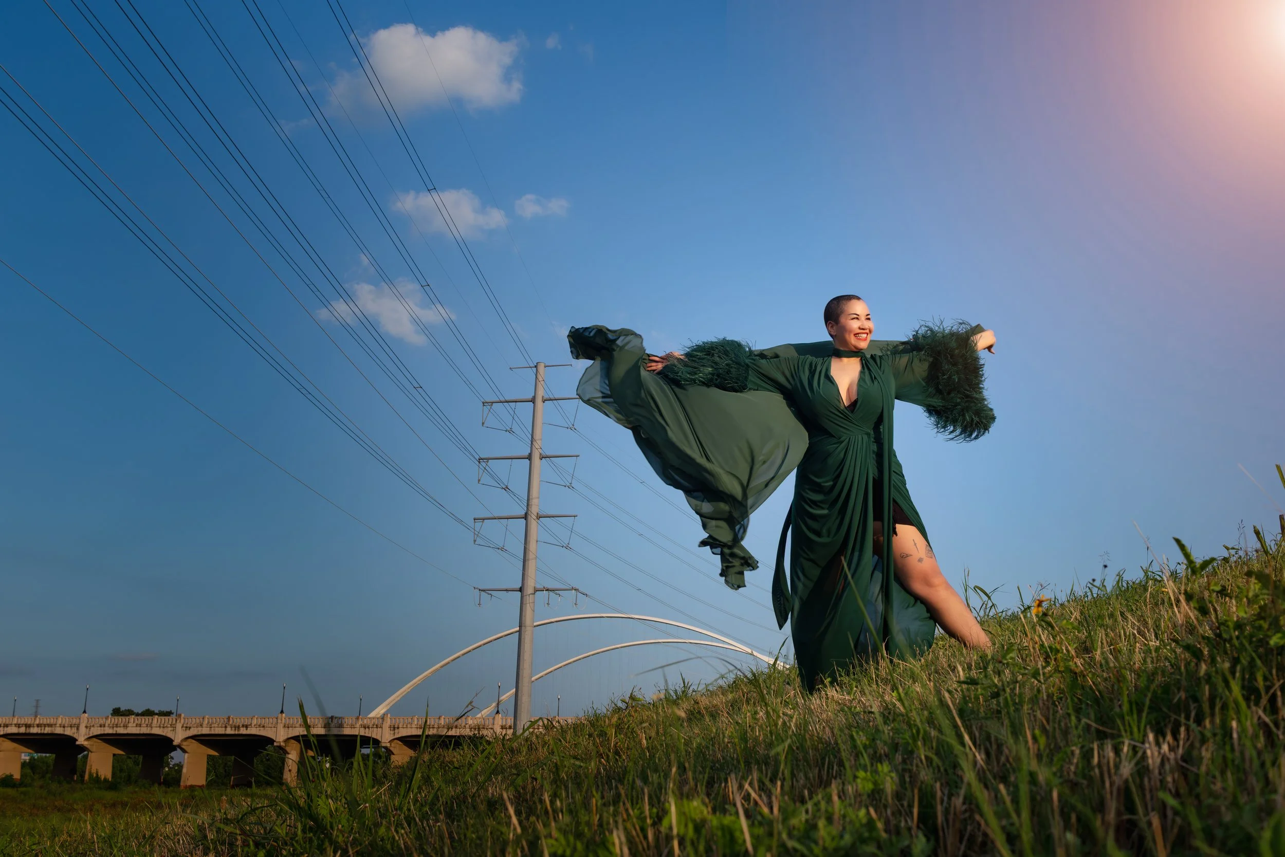 A person in a flowing green dress standing on a grassy hillside with arms outstretched, smiling, against a clear blue sky with power lines and a bridge in the background.