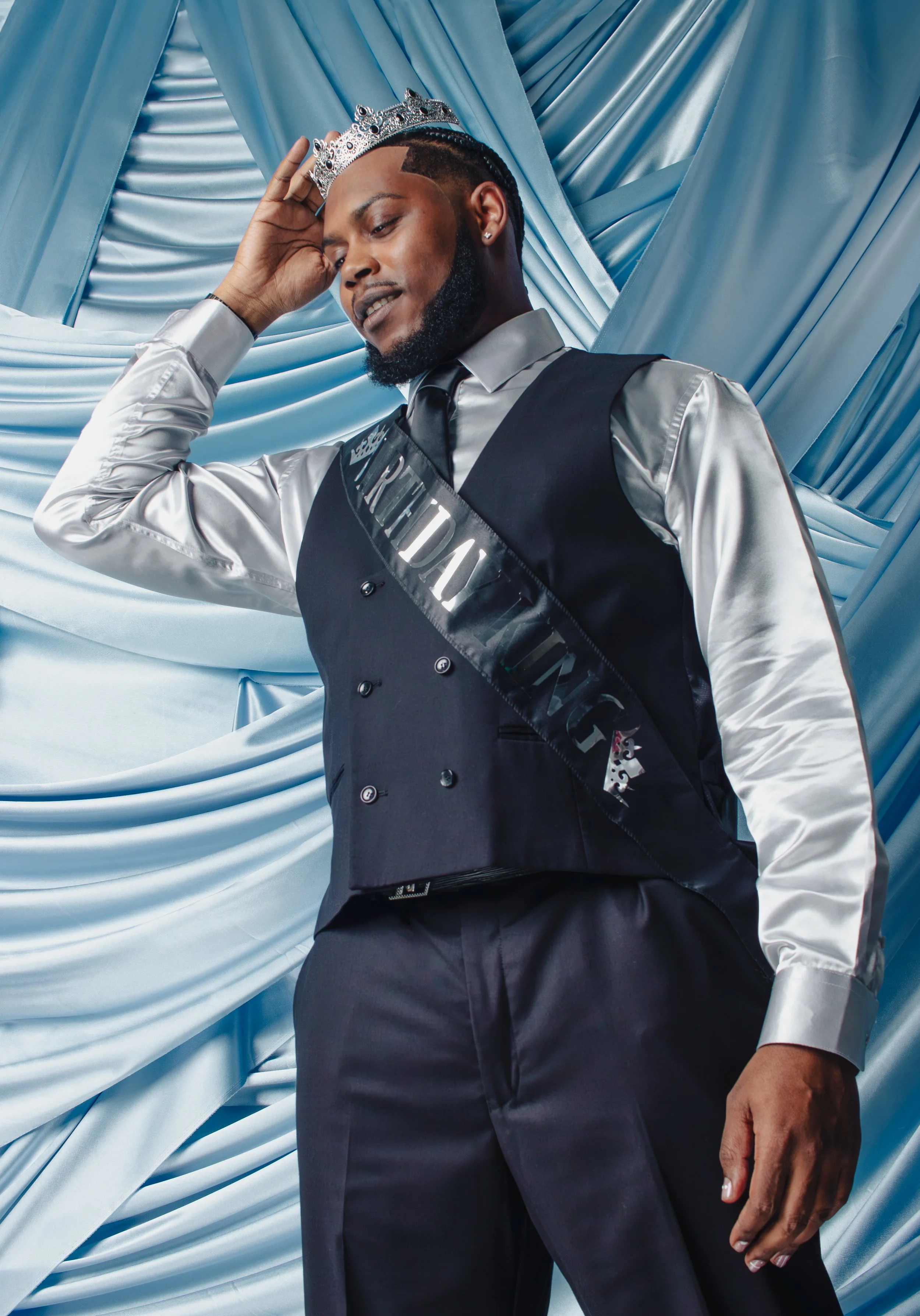 A man wearing a silver shirt, black vest, and black pants, with a crown and a sash that reads 'PARTY DUK,' is posing in front of a blue, satin-draped backdrop, with one hand touching his head.