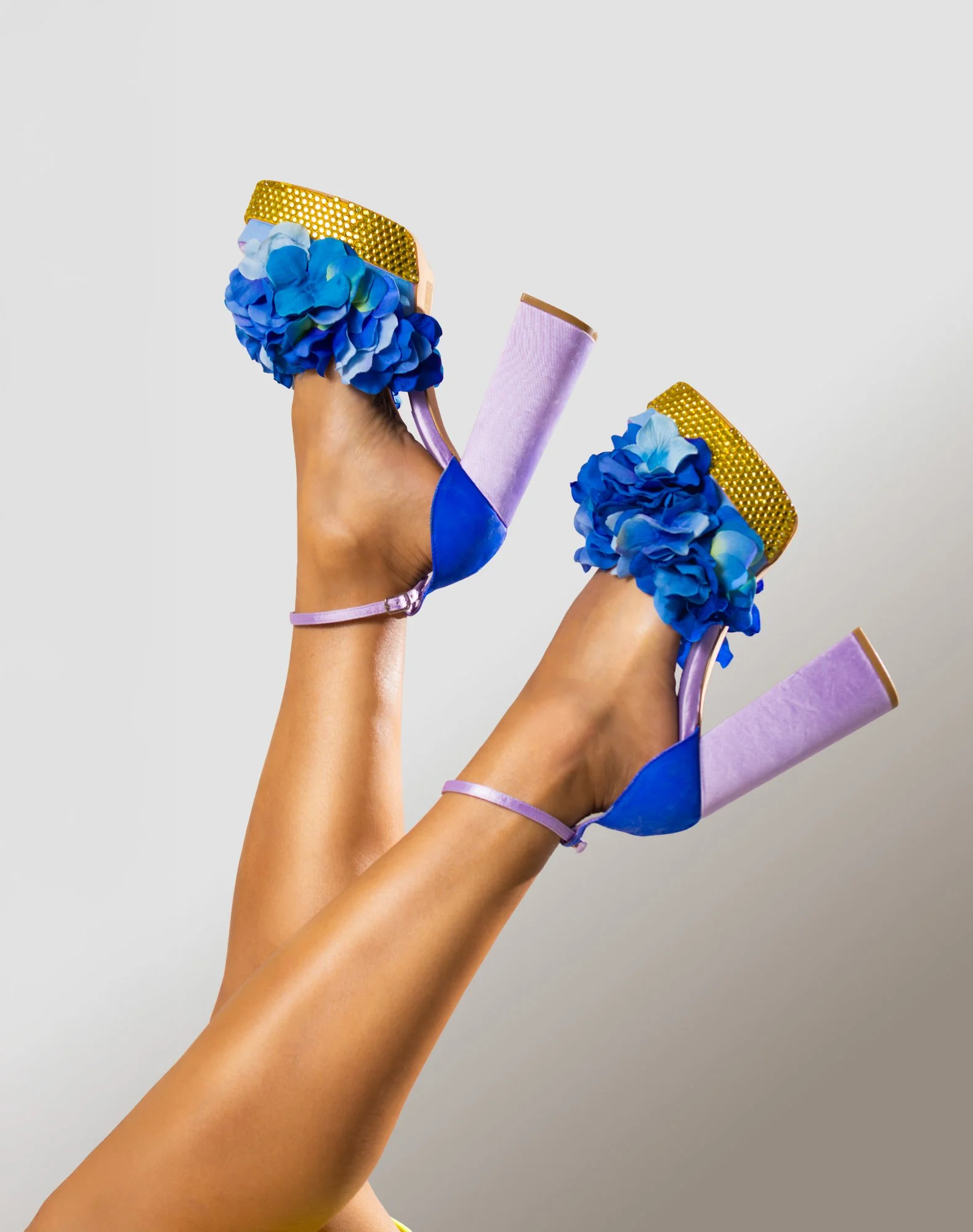 Pair of colorful high-heeled shoes with blue flowers, gold accents, and lavender heels, worn by a person with tanned legs raised against a plain background.