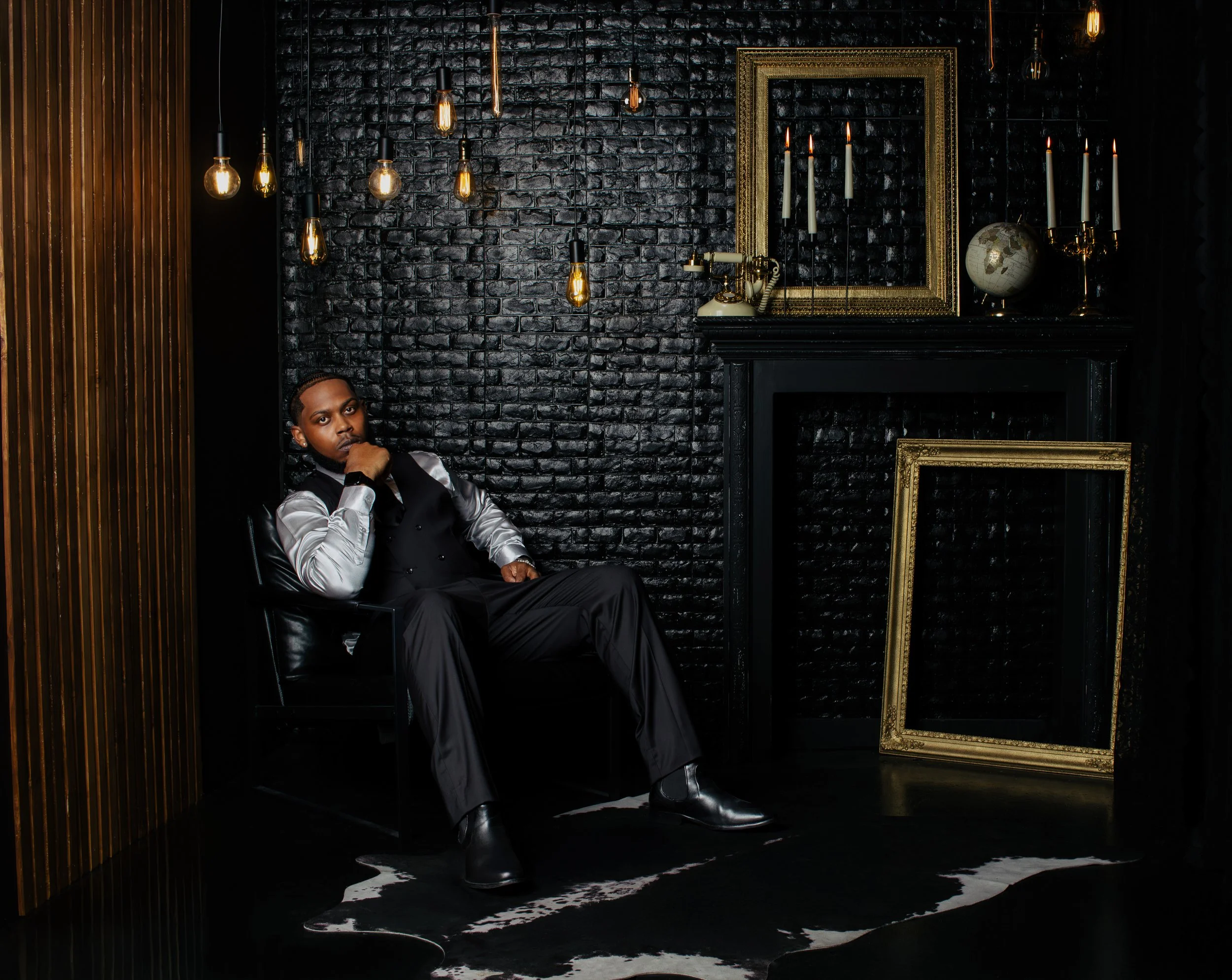 A man sitting in a black leather chair in a dark, modern room with black brick walls, gold framed mirrors, and Edison bulb hanging lights. The man is dressed in a black and silver outfit, with a thoughtful expression, and is resting his chin on his h