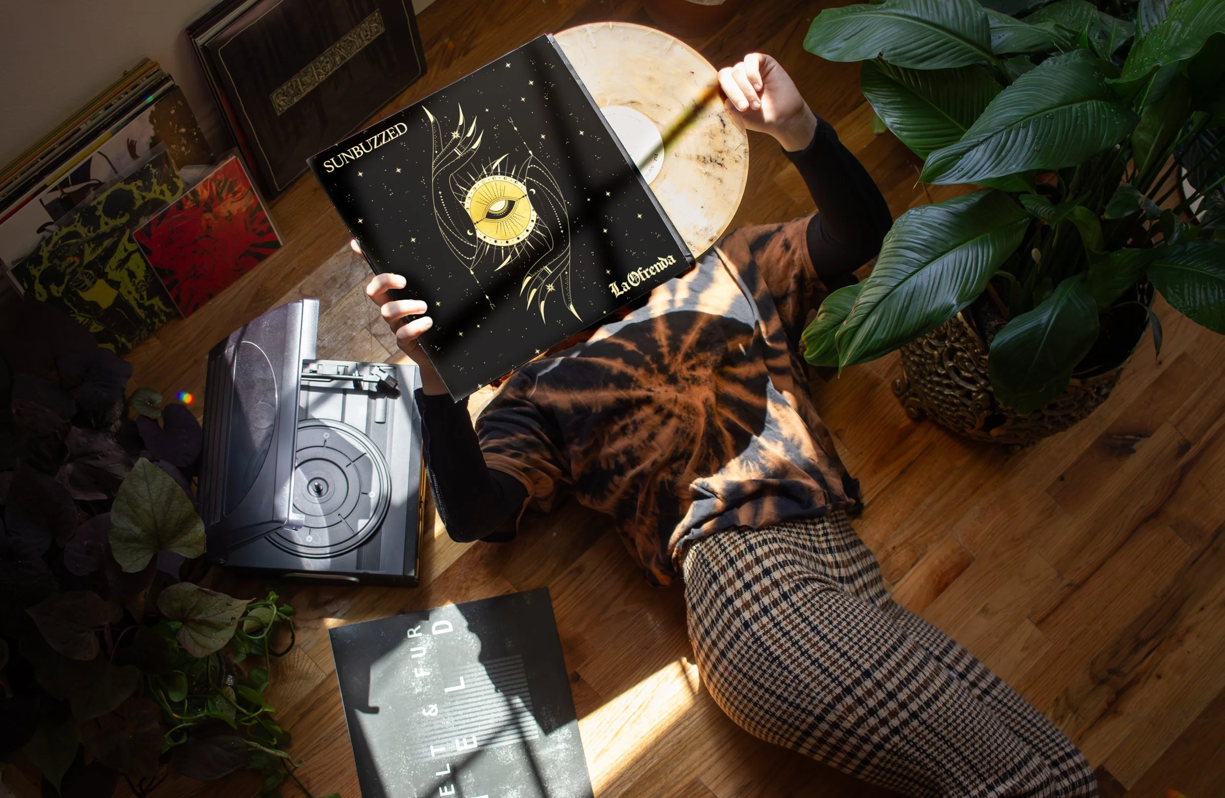 A person lying on the hardwood floor, holding a vinyl record album titled "Sunbuzzed" with a black cover featuring a golden eye and celestial design. The person is wearing checkered pants, a dark long-sleeved shirt, and a tie-dye t-shirt. Nearby are