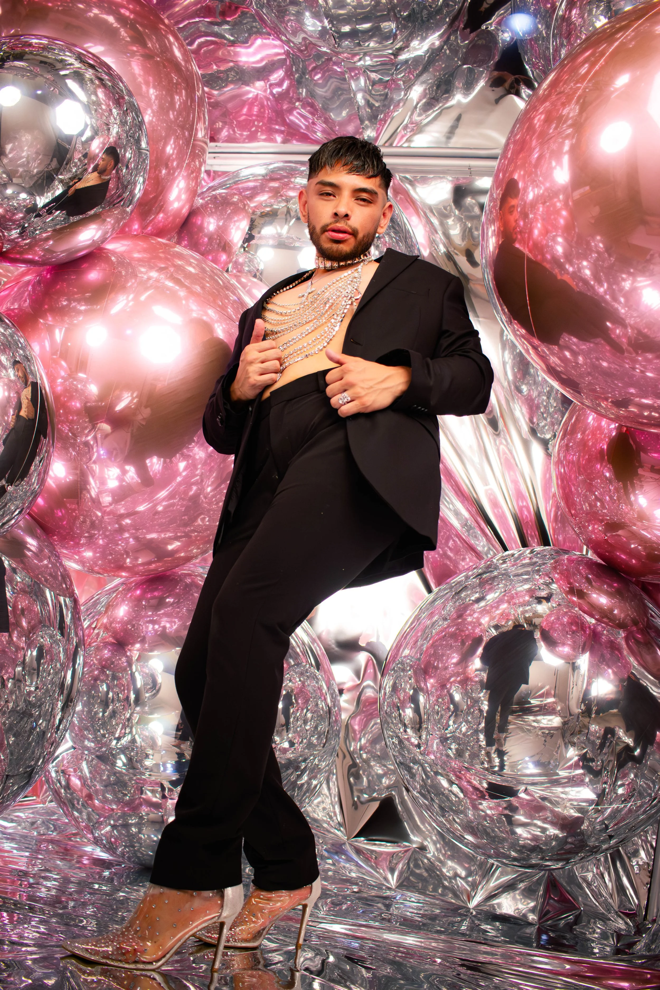 A person wearing a black suit, multiple silver and diamond necklaces, and transparent high heels with embellishments stands amidst reflective pink and silver balloons and metallic foil decorations.