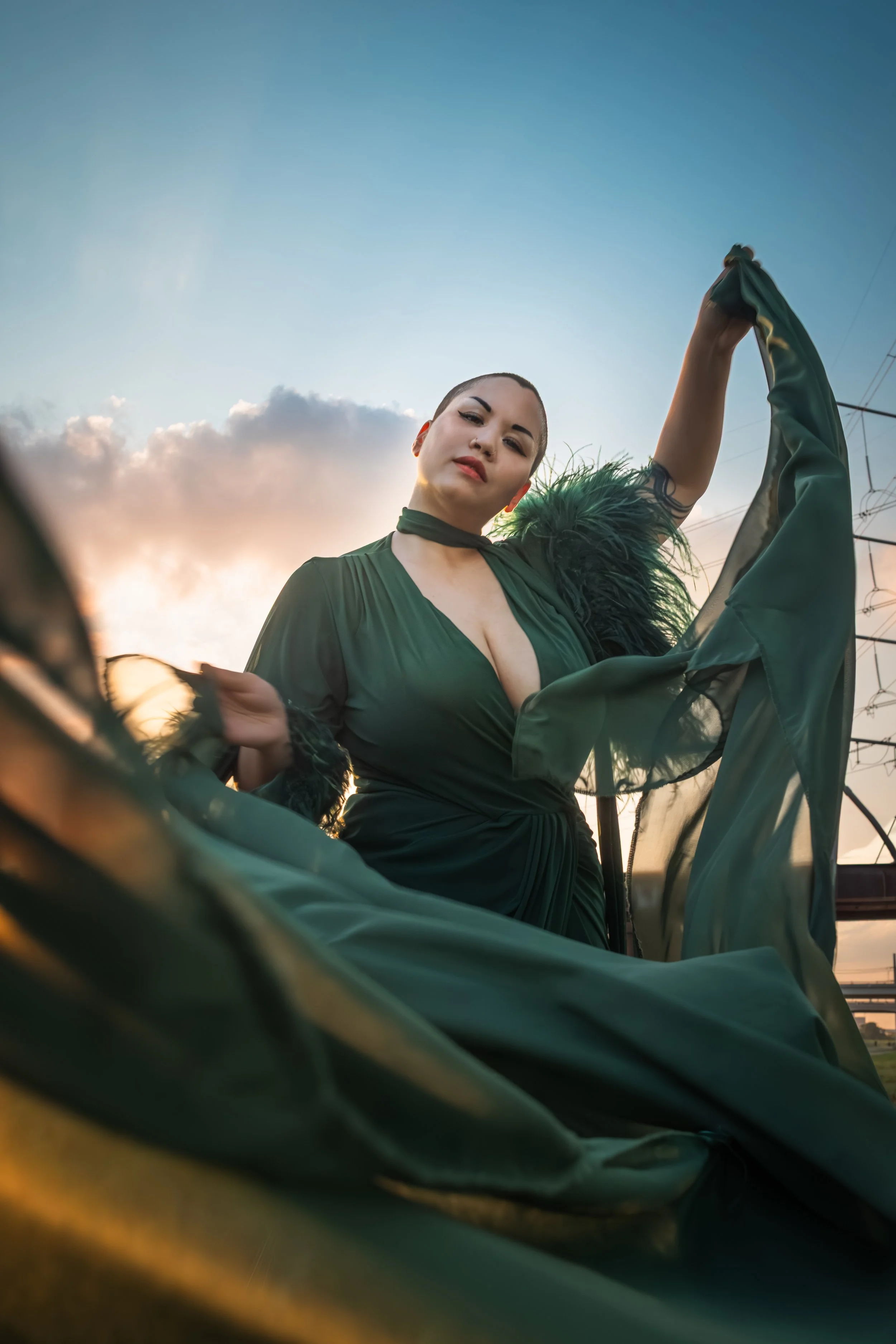 A person in a green dress holds up part of her dress outdoors during sunset, with power lines in the background.