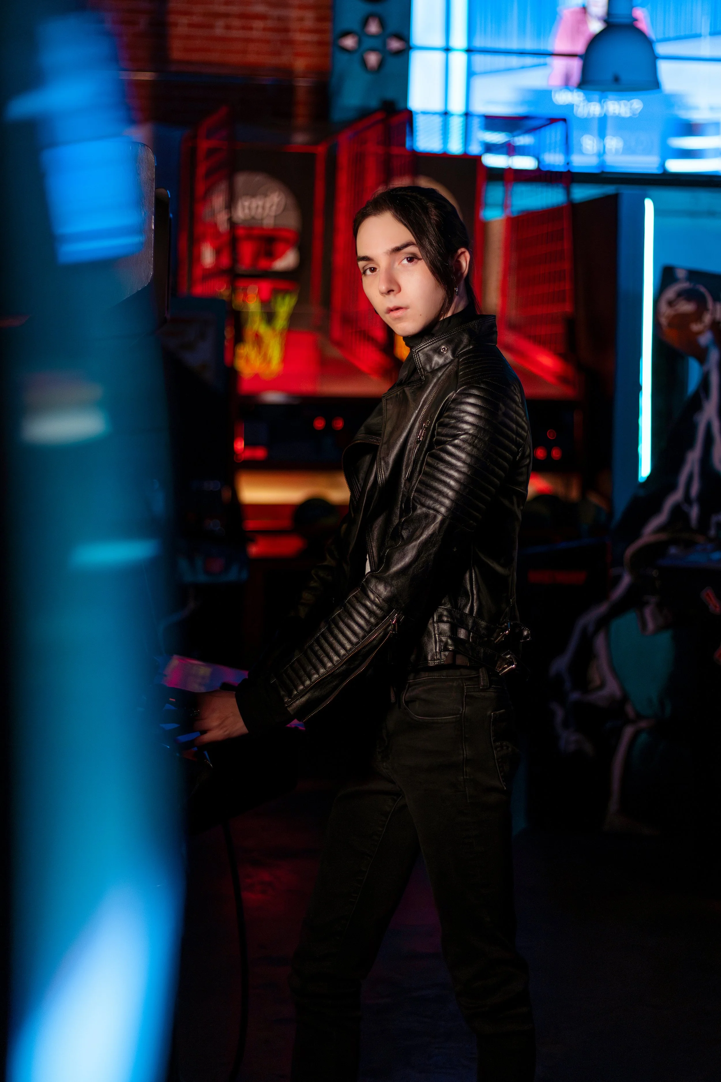 A young person dressed in black leather jacket and black pants in a dimly lit arcade, standing next to a gaming machine.