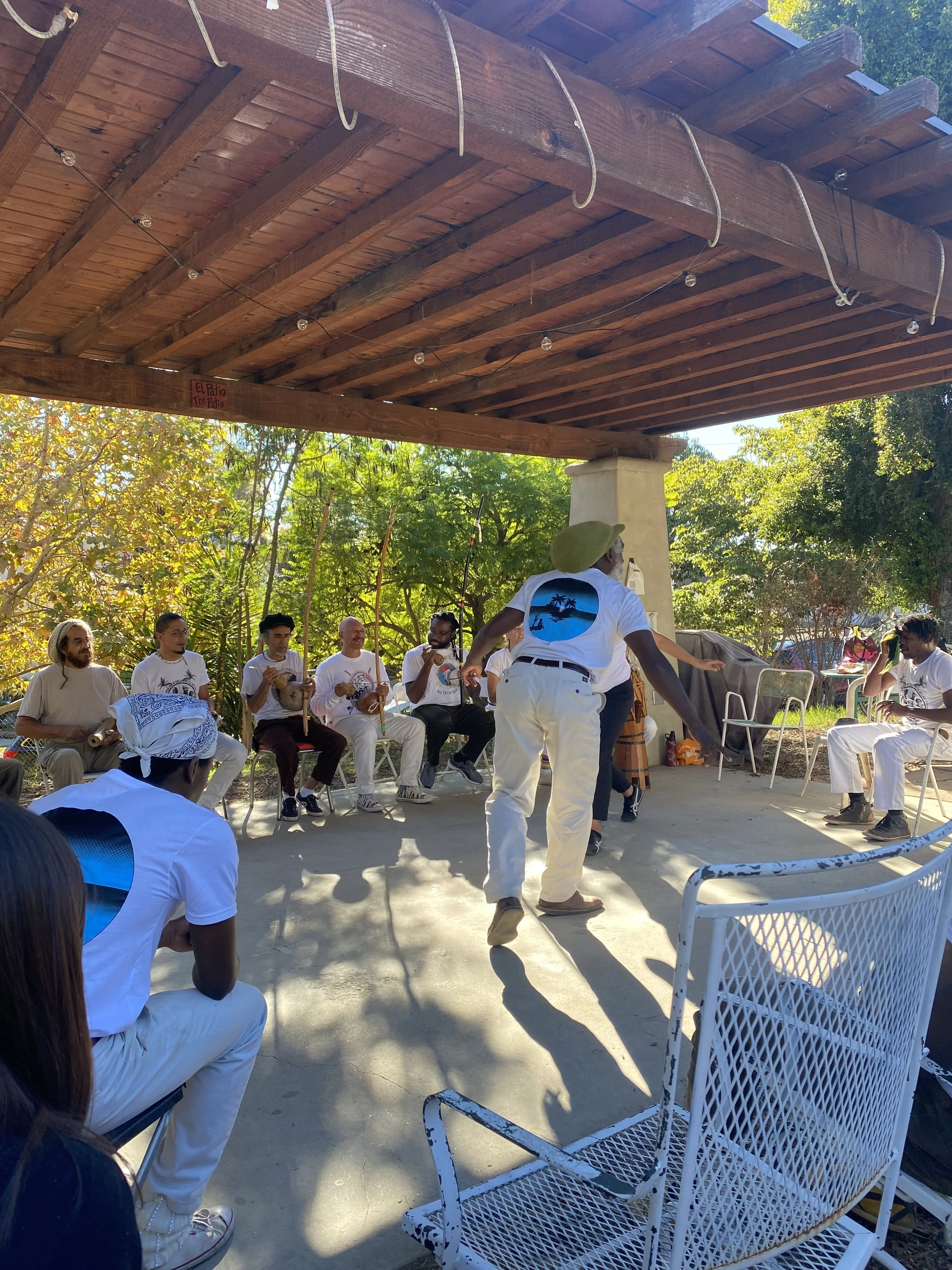 A group of people sitting in a semi-circle under a wooden canopy, participating in a Capoeira de Angola cultural music and martial art display outdoors on a sunny day in Southern California during an N'gola Preto Velho roda.