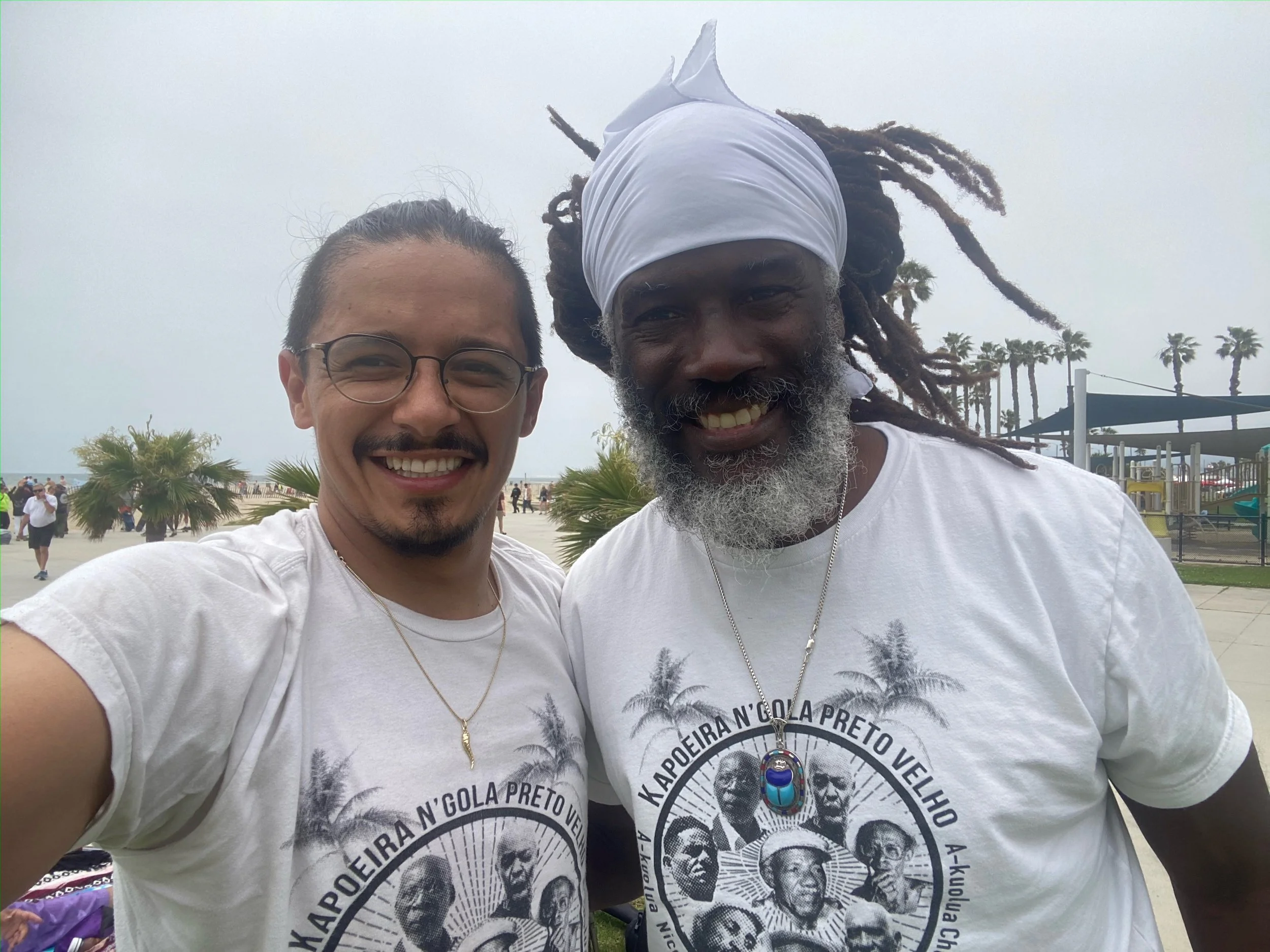 Capoeira de Angola Instructor Nico Zuluaga and Treinel Mo smile and take a selfie outdoors. Both wearing N'gola Preto Velho T-shirts. Background shows a park with palm trees and a cloudy sky.