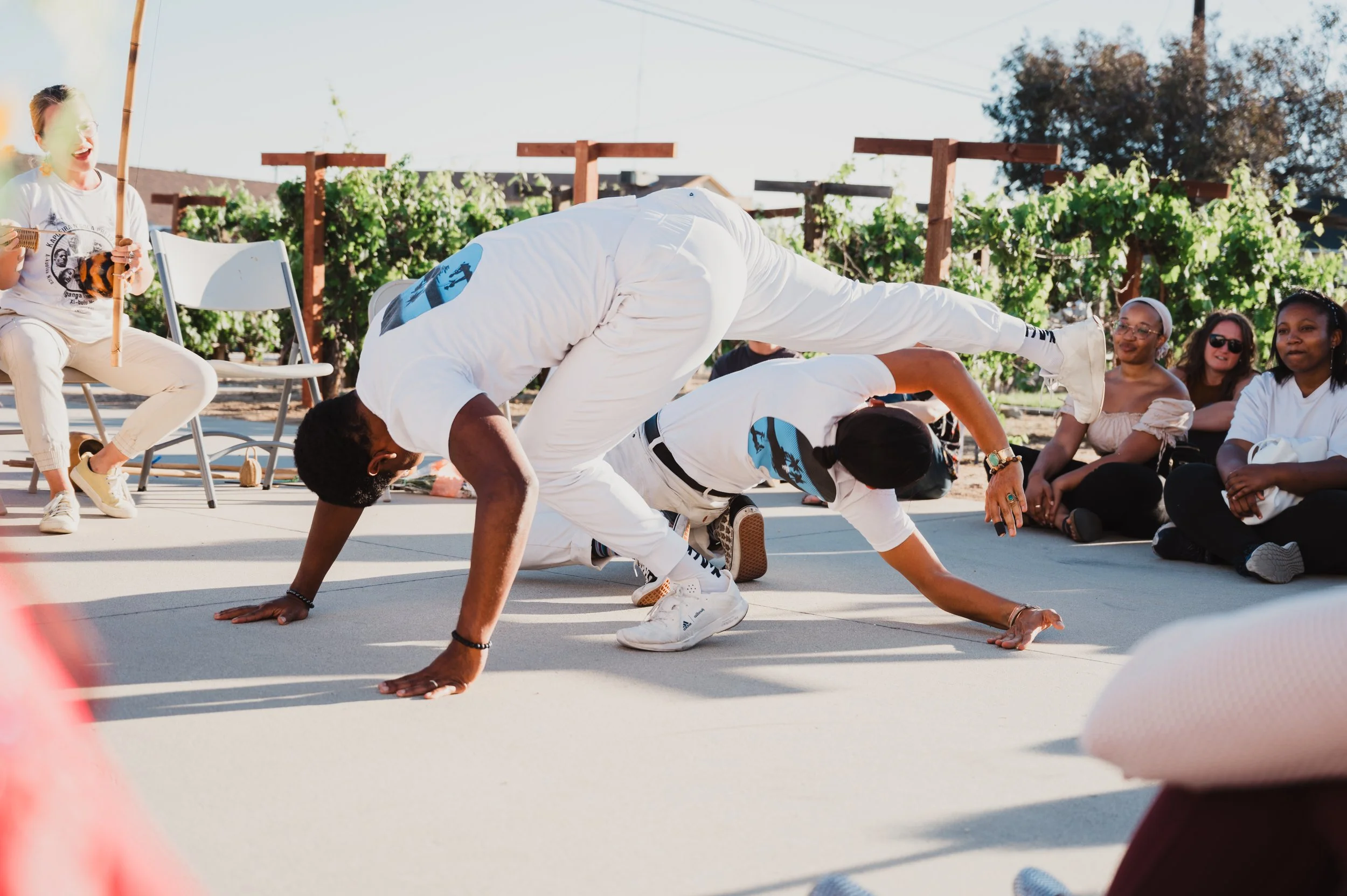Capoeira de Angola instructor Nico Zuluaga and student of N'gola Preto Velho engage in a capoeira presentation in Southern California. The scene is outdoors with greenery in the background.