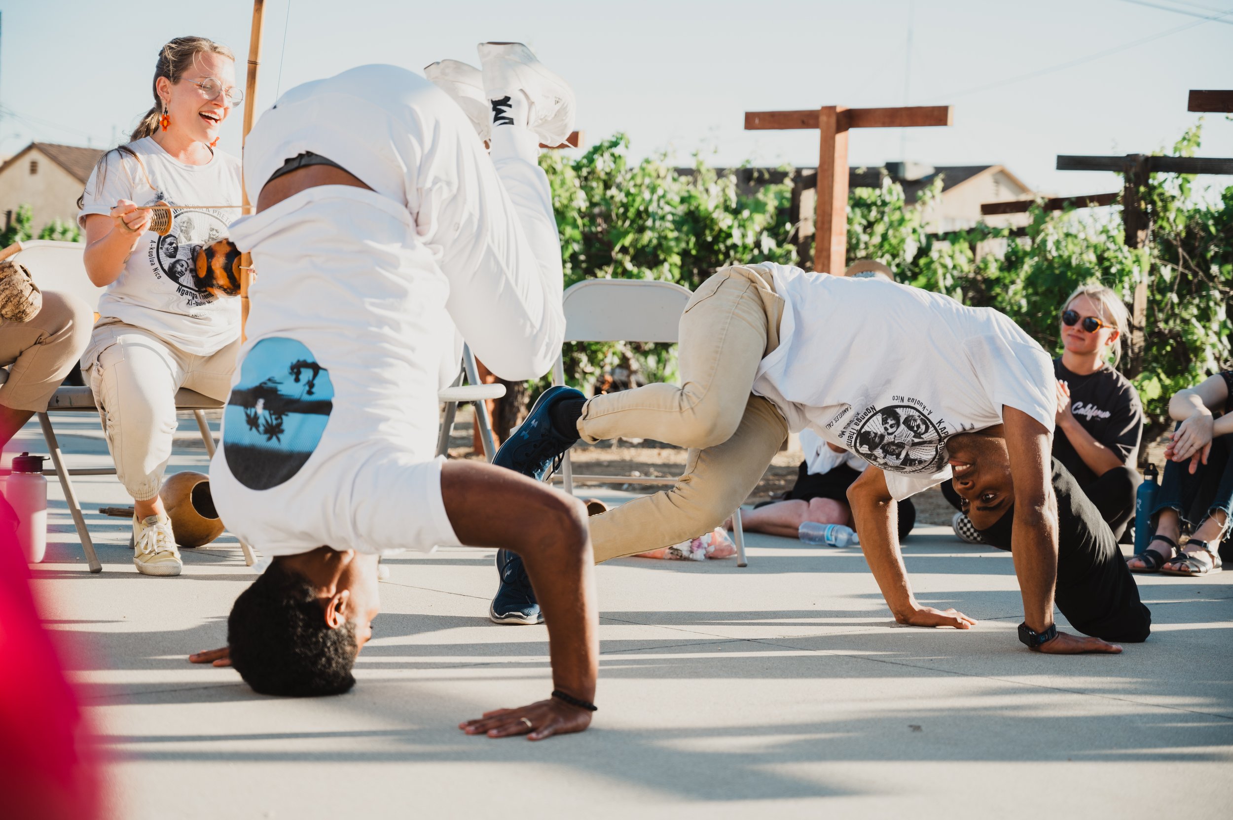 People observe an outdoor performance organized by N'gola Preto Velho, a Capoeira de Angola school in Southern California, with each person displaying an Au de Cabeça headstand while others sit and cheer in the background.
