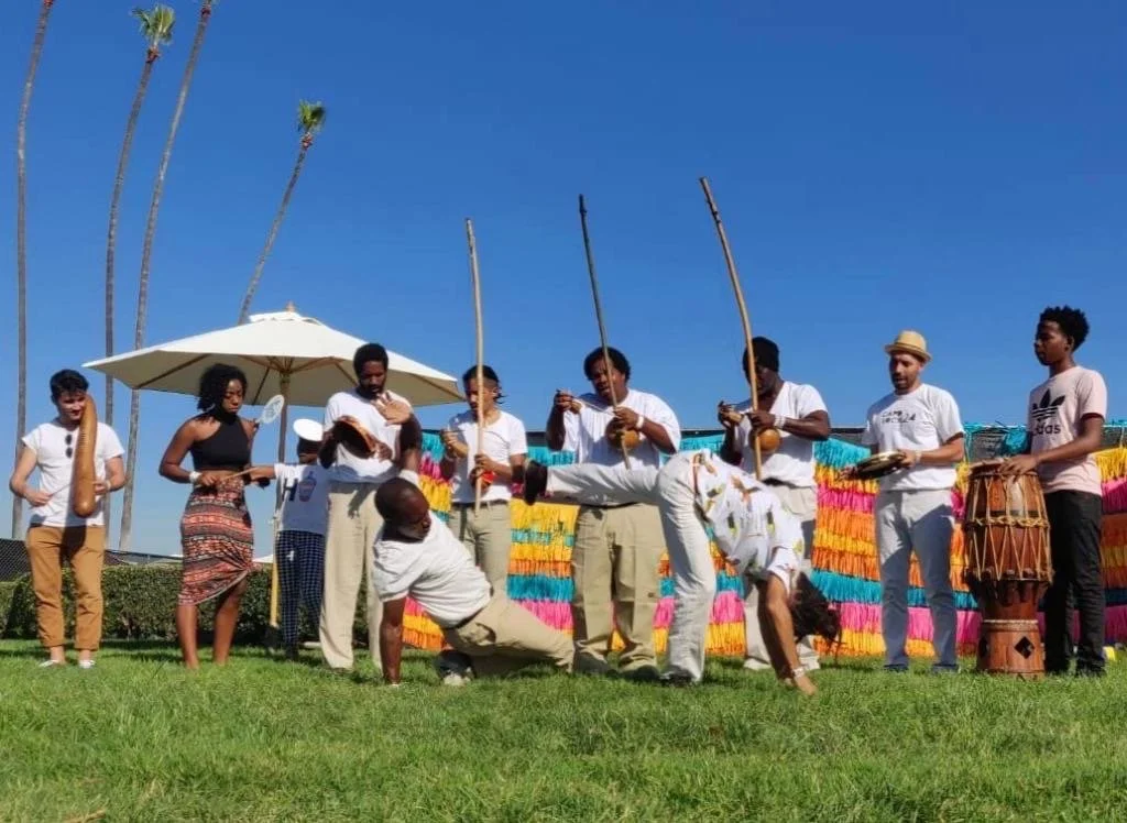 Group of people performing a the Afro-Brazilian martial art Capoeira de Angola during an outdoor performance in Southern California, under a blue sky with palm trees.