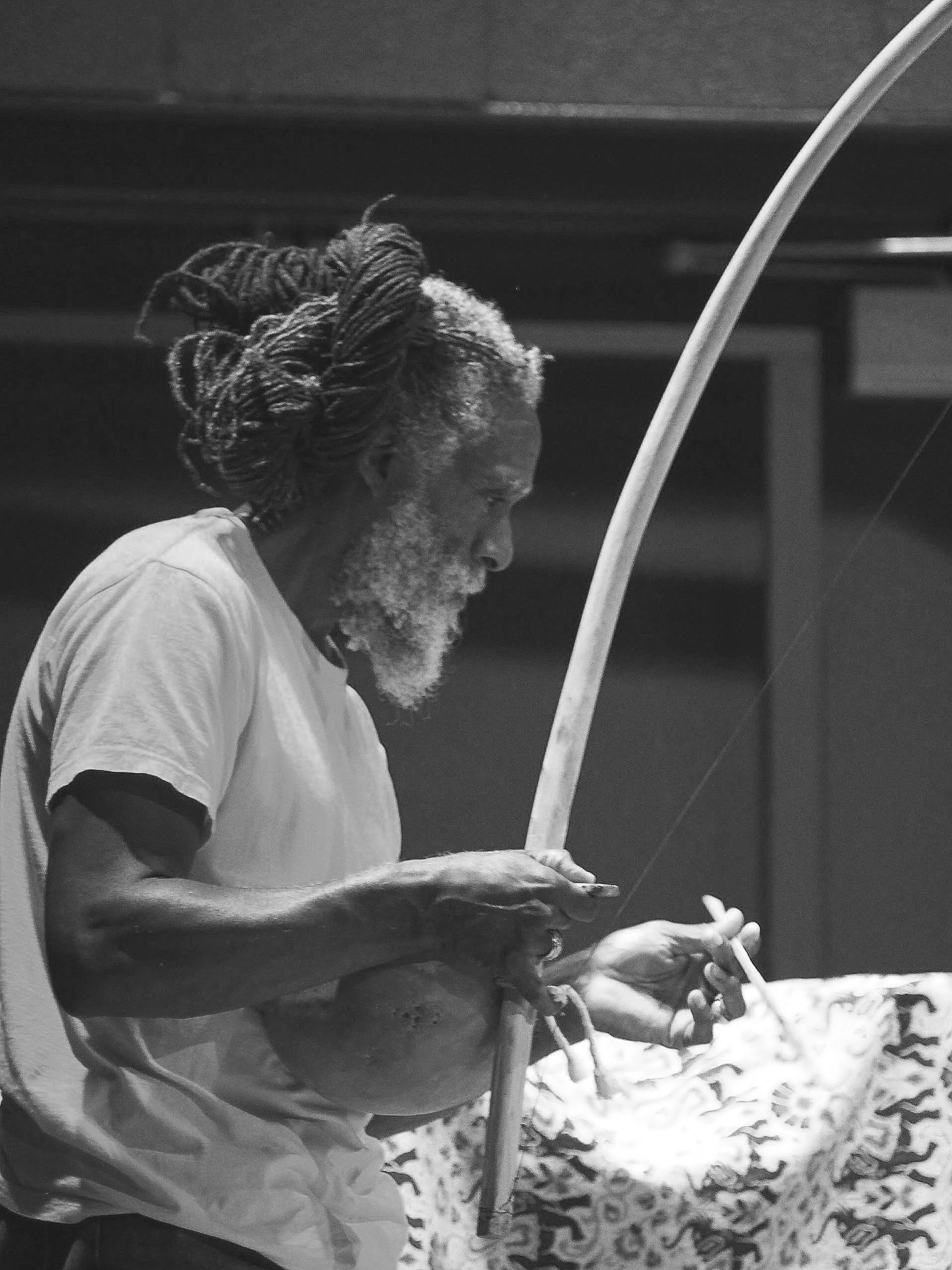 A black and white photo of an elderly man with long, dreadlocked hair and a beard, wearing a t-shirt, holding a fishing rod and a cigarette.