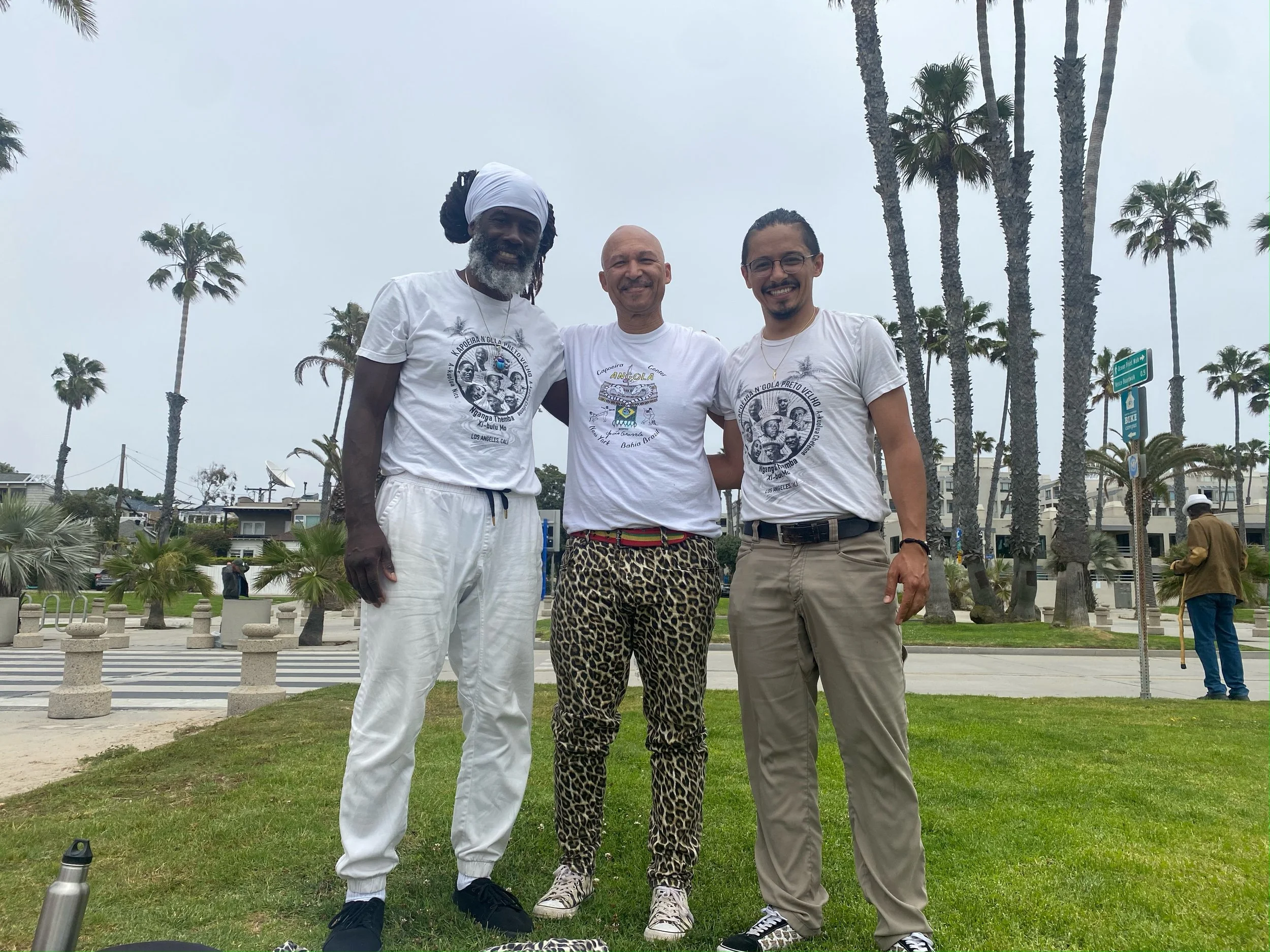 Members of N'gola Preto Velho standing on a grassy area with palm trees in the background, smiling and posing for a photo during a cloudy day after a Capoeira de Angola roda and performance. 