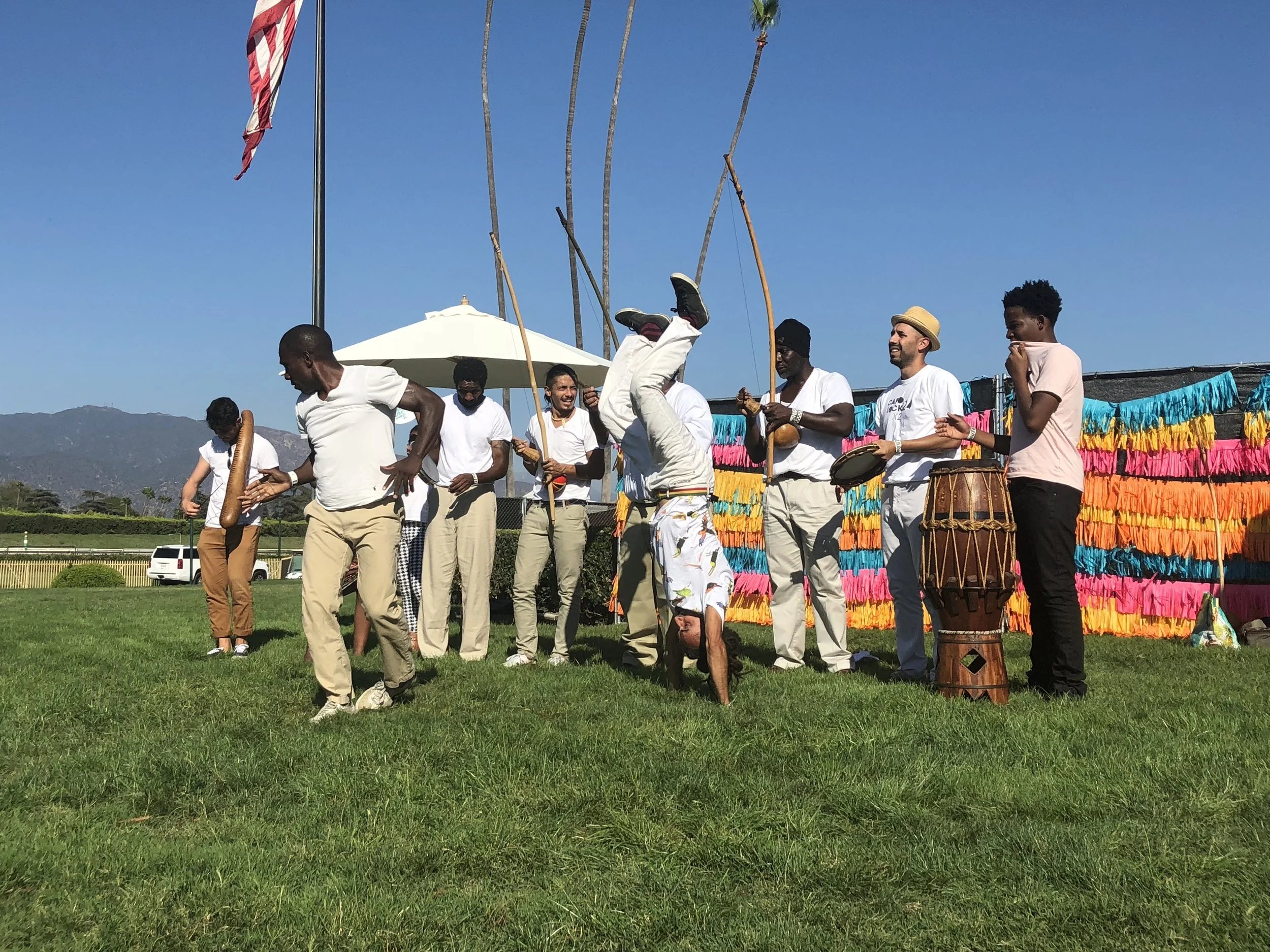 Group of people play Capoeira de Angola outdoors with colorful decorations and a flag in the background.
