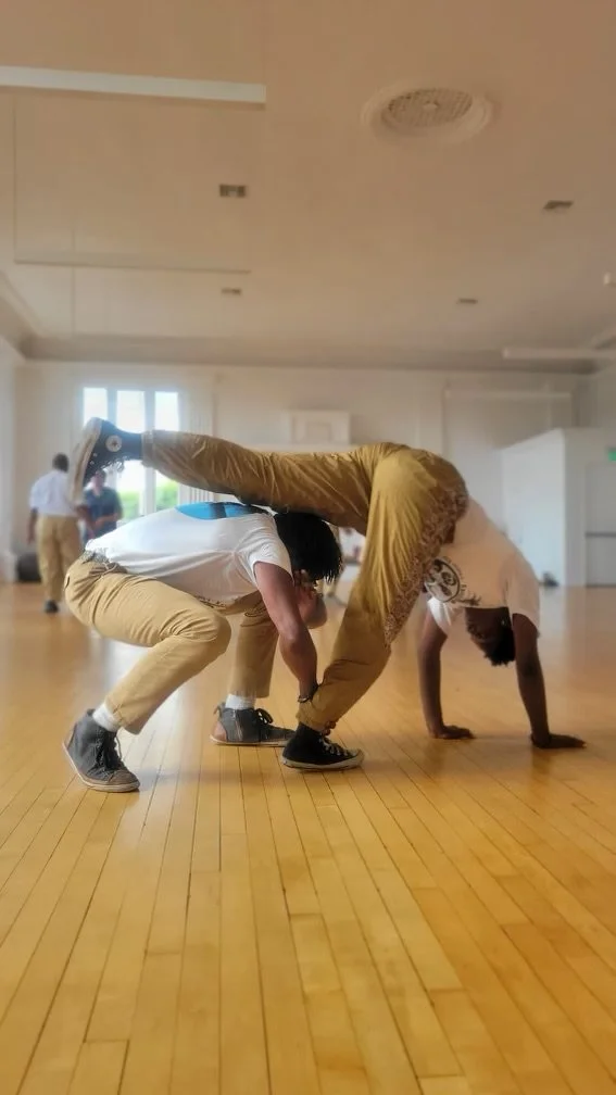 Members of N'gola Preto Velho practice Capoeira de Angola in a performance call with natural light coming through windows. One person is doing a handstand or Au while the other two support their body in a balanced manner.