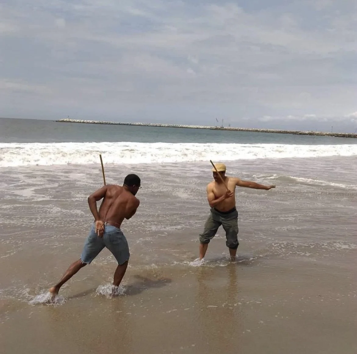 Two men, shirtless and wearing shorts, are playing Capoeira with sticks in the shallow surf at a beach, with a breakwater and cloudy sky in the background.