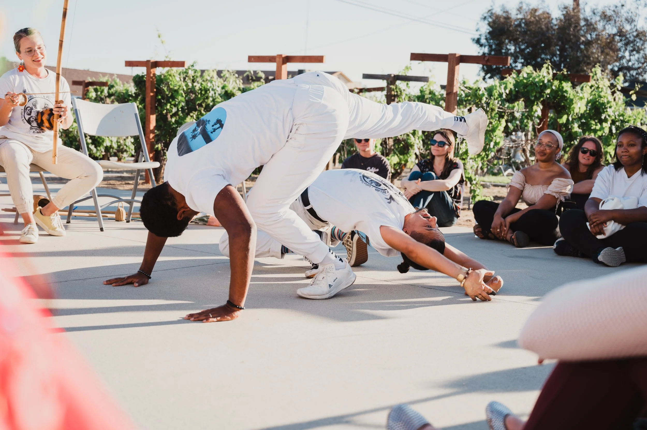 Capoeira de Angola instructor Nico Zuluaga and student engage in a Capoeira de Angola performance while an audience watches, outdoors with sunlight and greenery in the background in Southern California.
