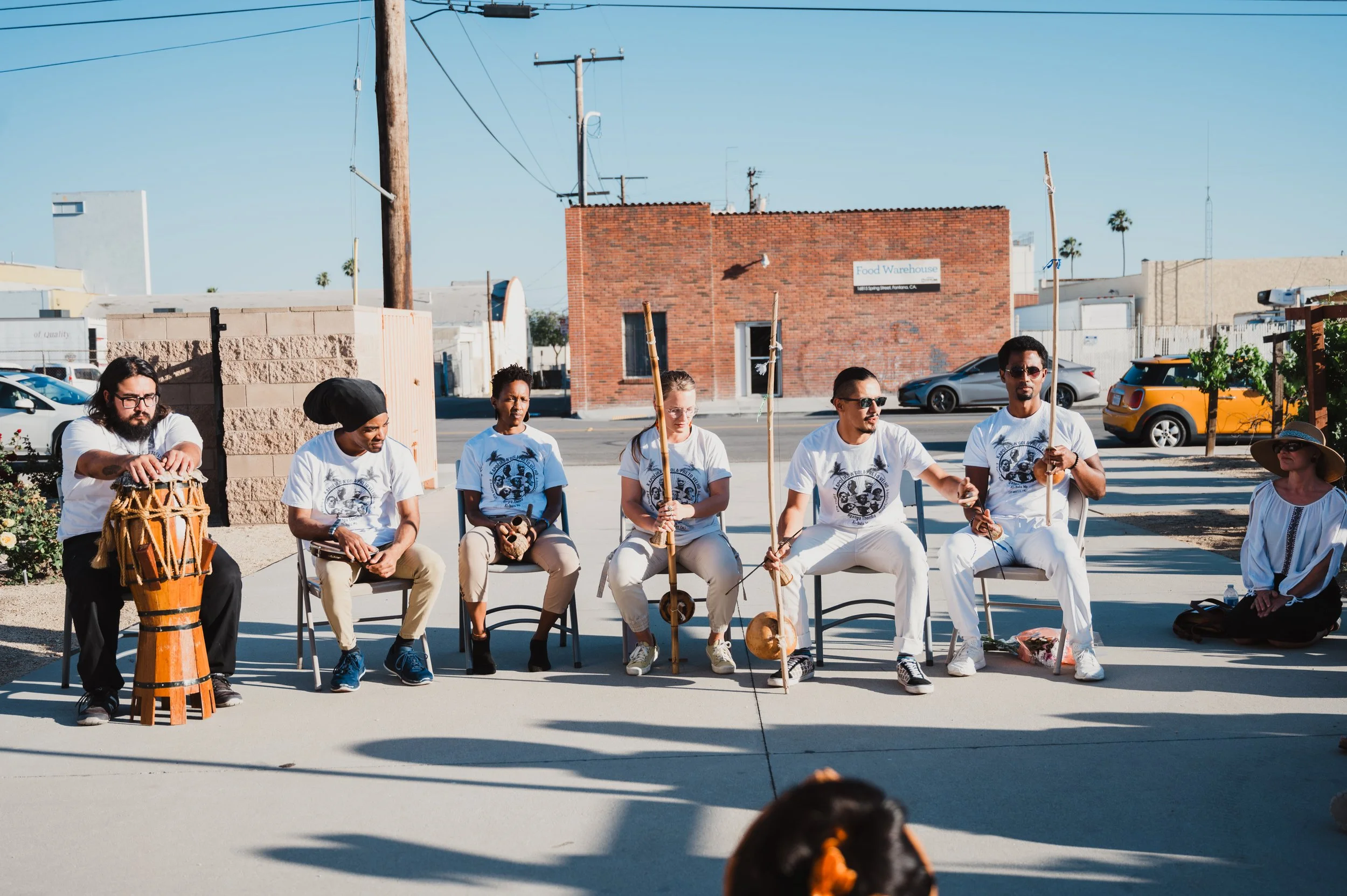Members of N'gola Preto Velho sit in a capoeira batería outdoors, playing traditional capoeira music, under a clear blue sky, with buildings and parked cars in the background.